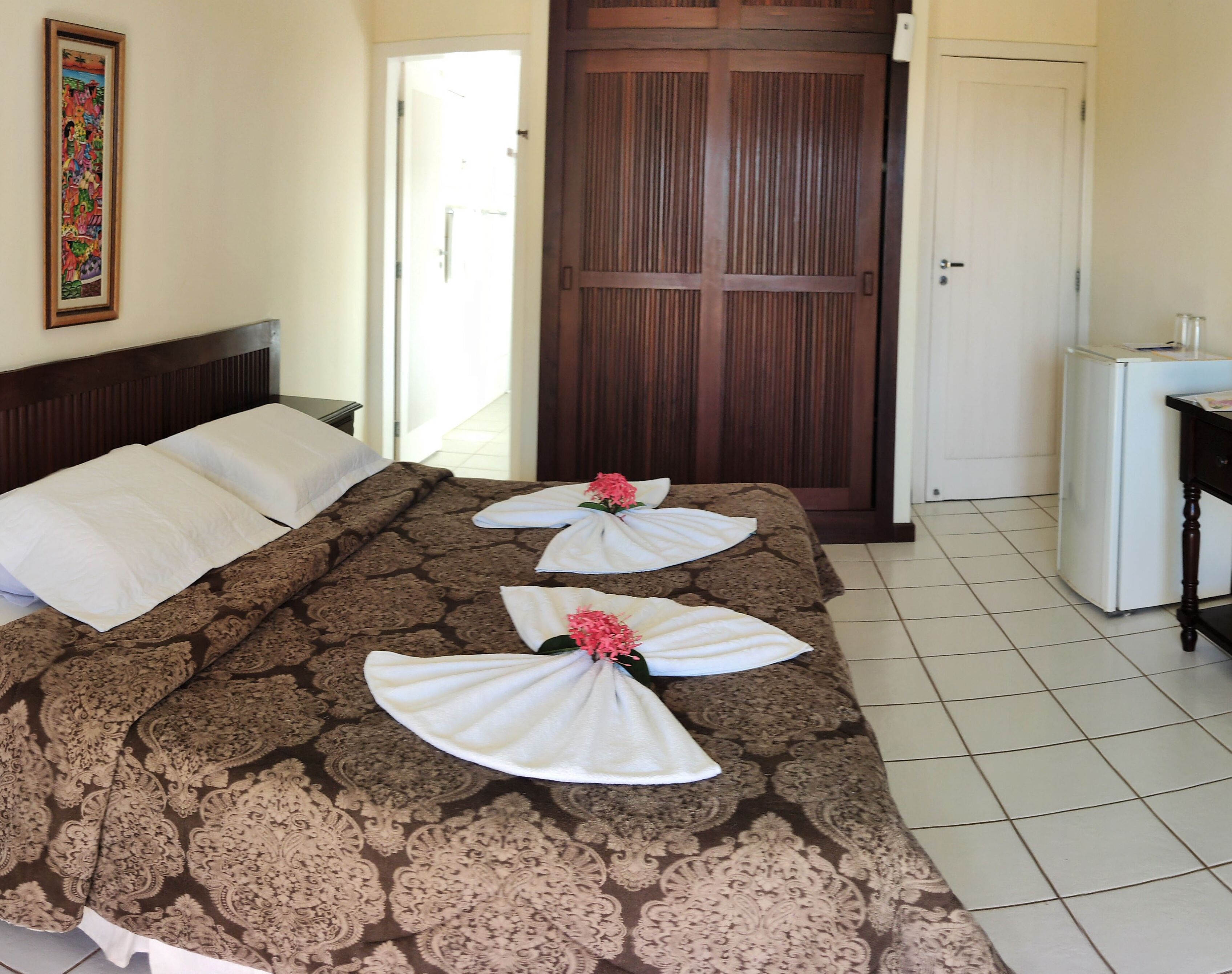 Quarto de hotel com uma cama grande, porta de madeira, pequena escrivaninha e geladeira.