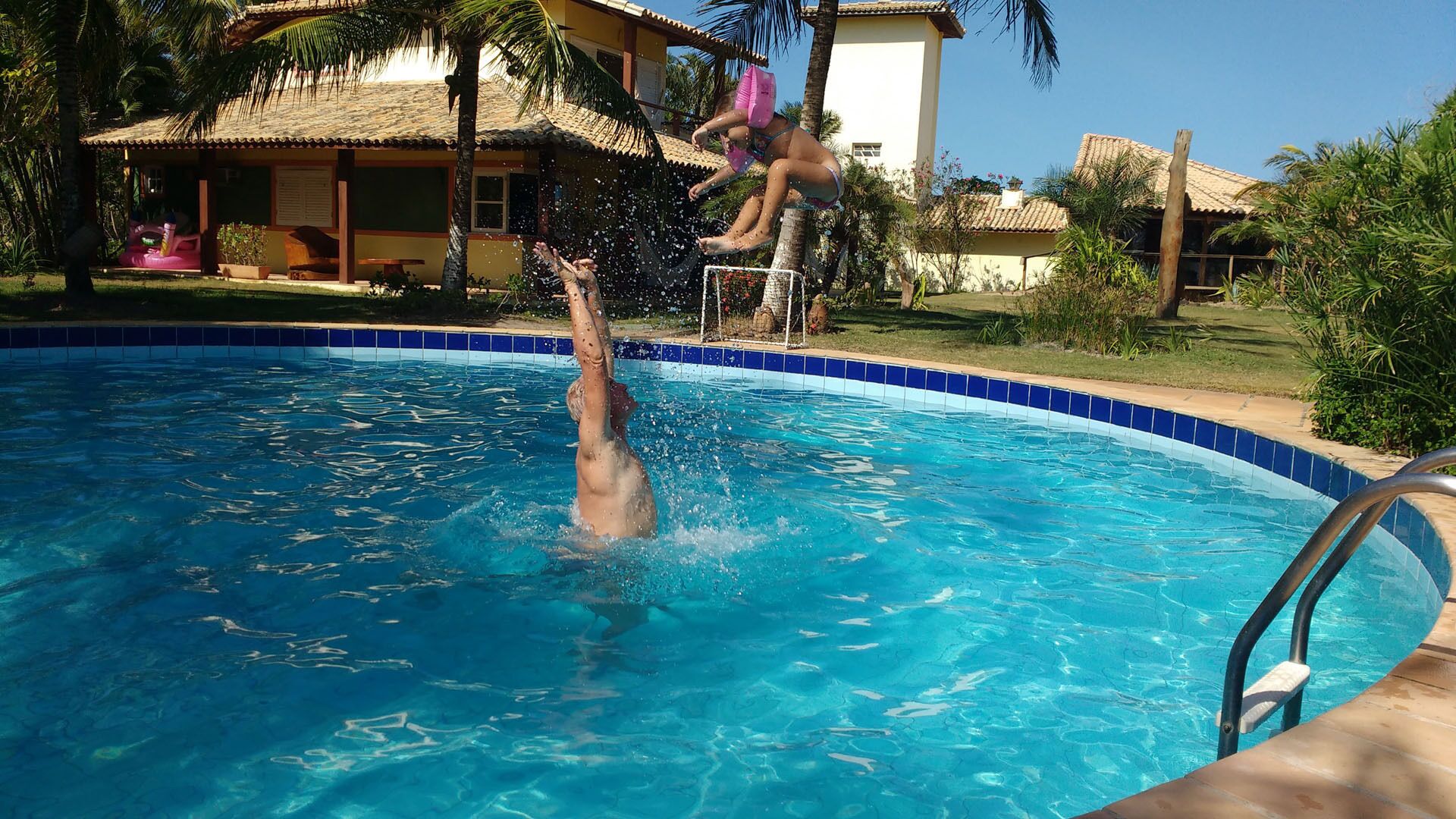En swimmingpool med krystalklart blåt vand, omgivet af et tropisk miljø med palmer og en bygning med et tag af fliser.