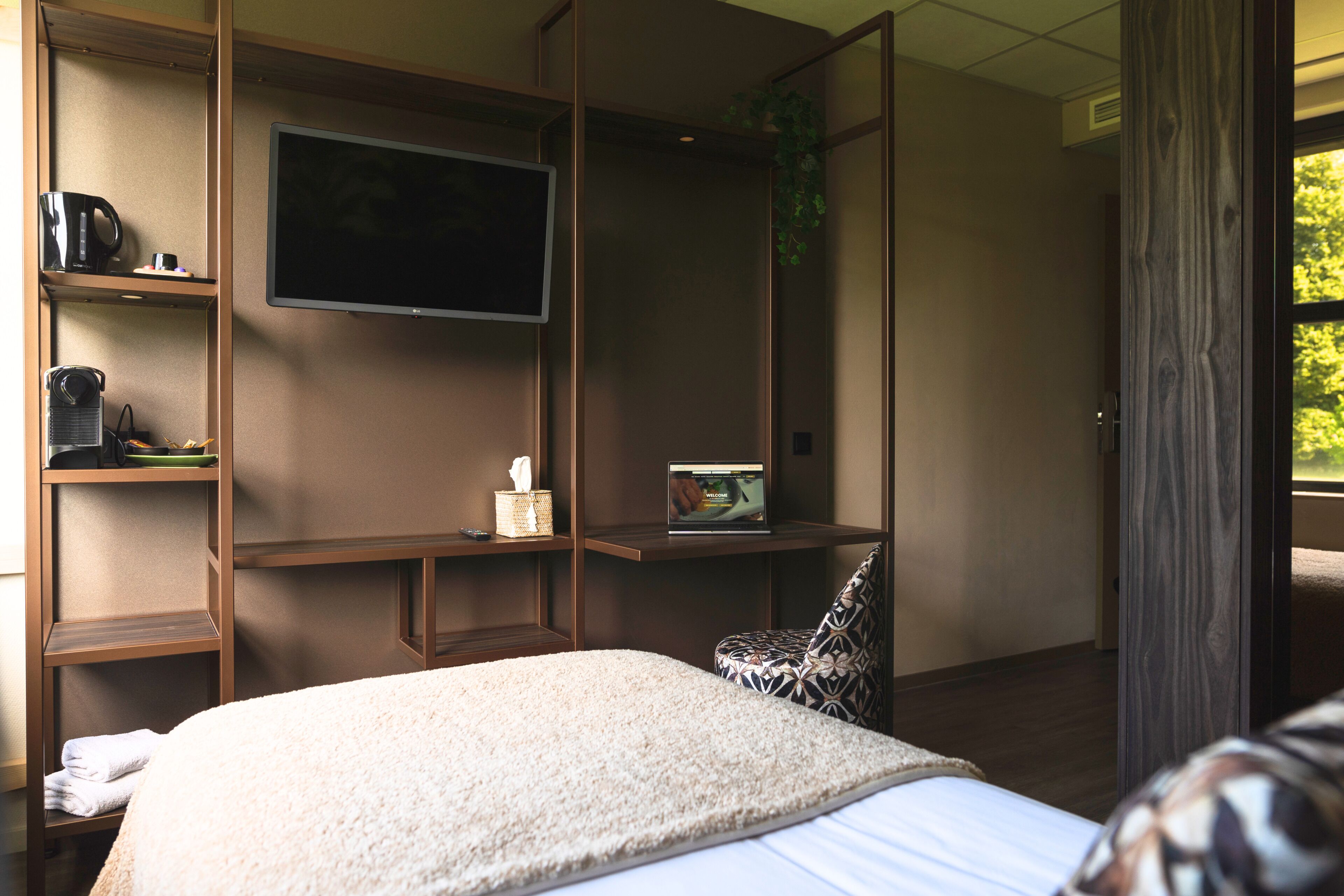 Una habitación de hotel con cama, televisión, escritorio con una computadora portátil, cafetera y ventana con vistas.