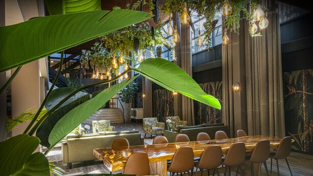 A dining area with a long wooden table, multiple chairs, and a backdrop of large green plants and hanging light fixtures. - Hotel Wander (Wageningen)