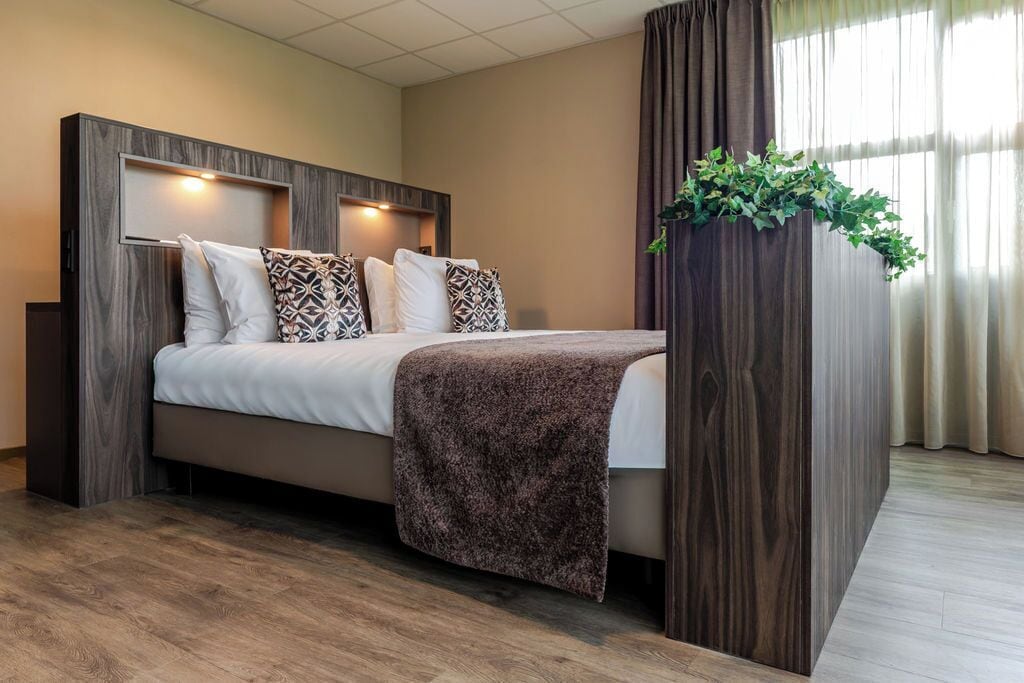 Ein modernes Hotelzimmer mit einem Bett, einem hölzernen Kopfteil, einer Pflanze und einem Fenster mit Vorhängen.