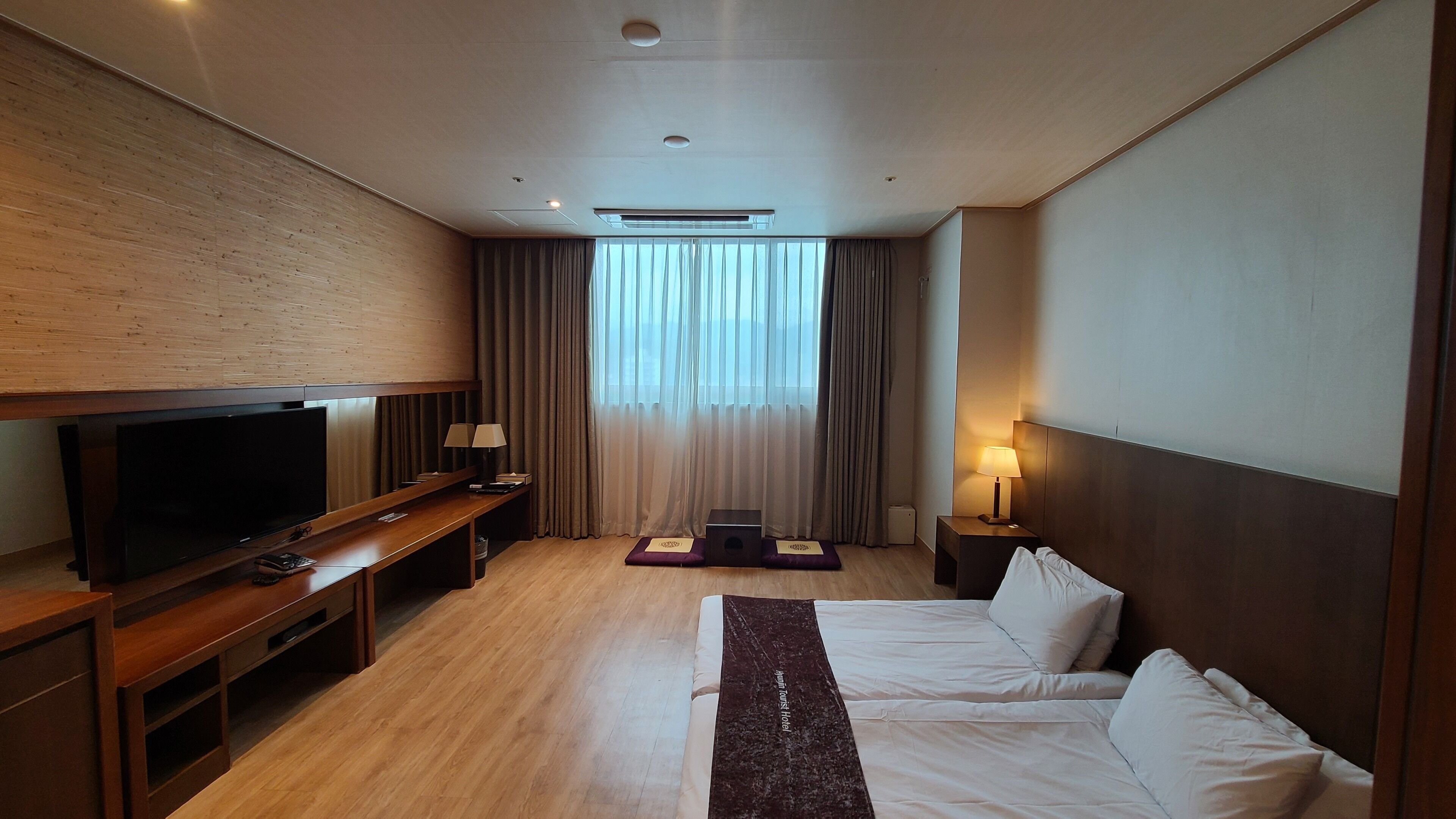 A hotel room with a bed, a TV, a desk, a lamp, and a window with curtains.
