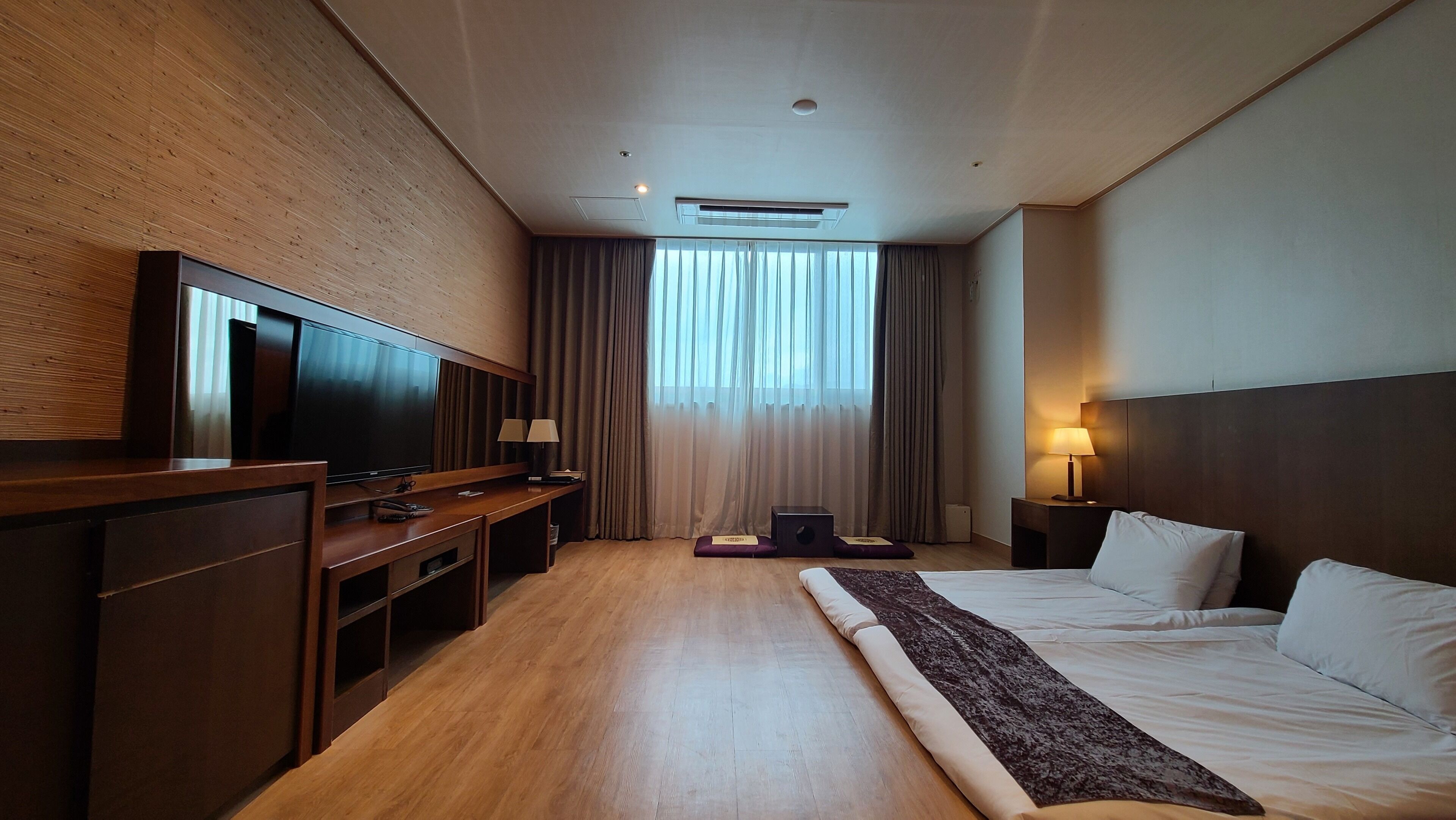 A hotel room with a large flat-screen TV, a wooden desk, a bed with white linens, a standing lamp, and a window with curtains.