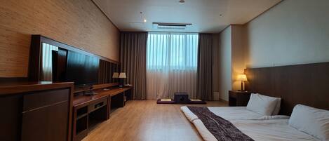 A hotel room with a large flat-screen TV, a wooden desk, a bed with white linens, a standing lamp, and a window with curtains.