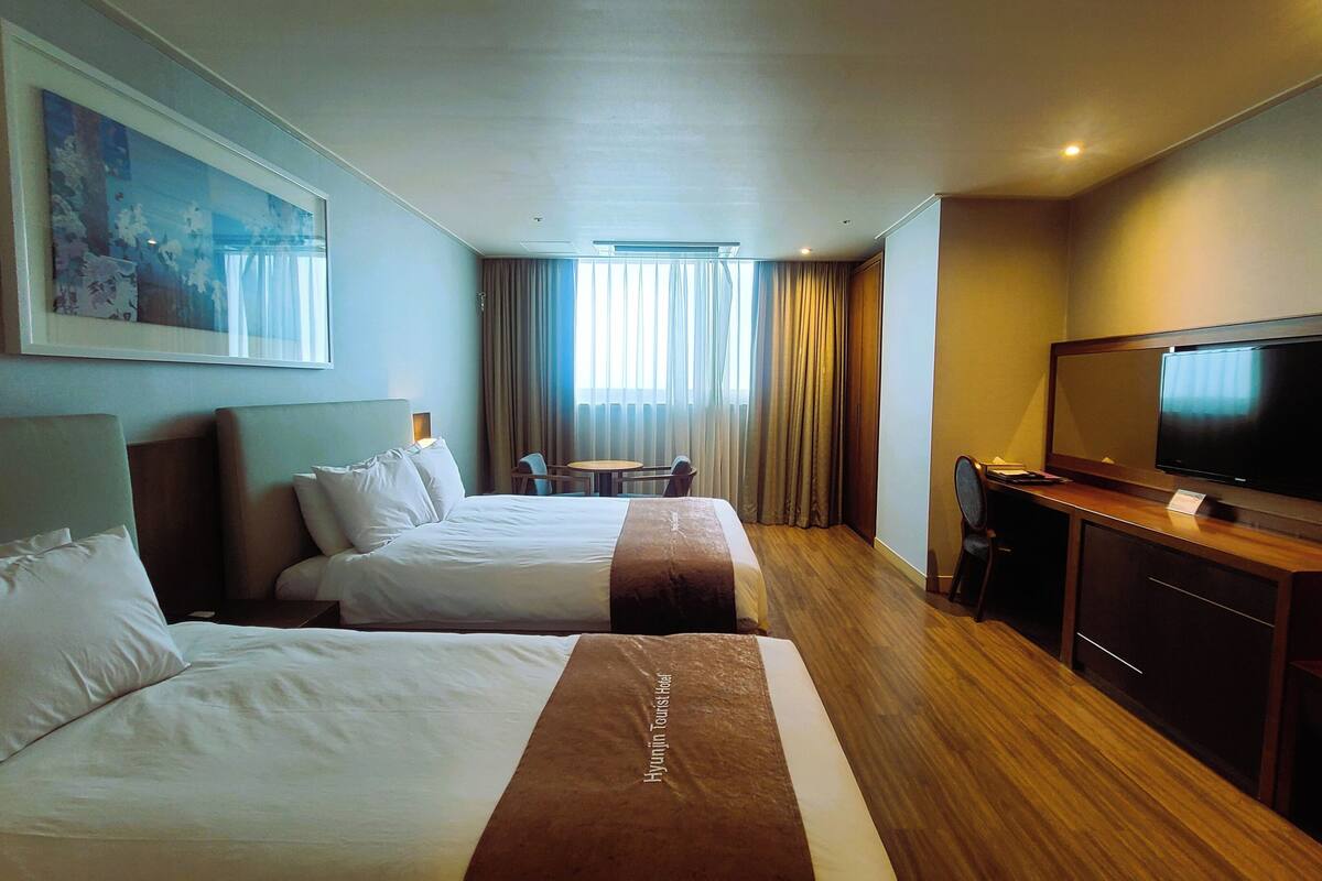 Habitación de hotel con dos camas, un escritorio con televisor, un cuadro en la pared y una ventana con cortinas.