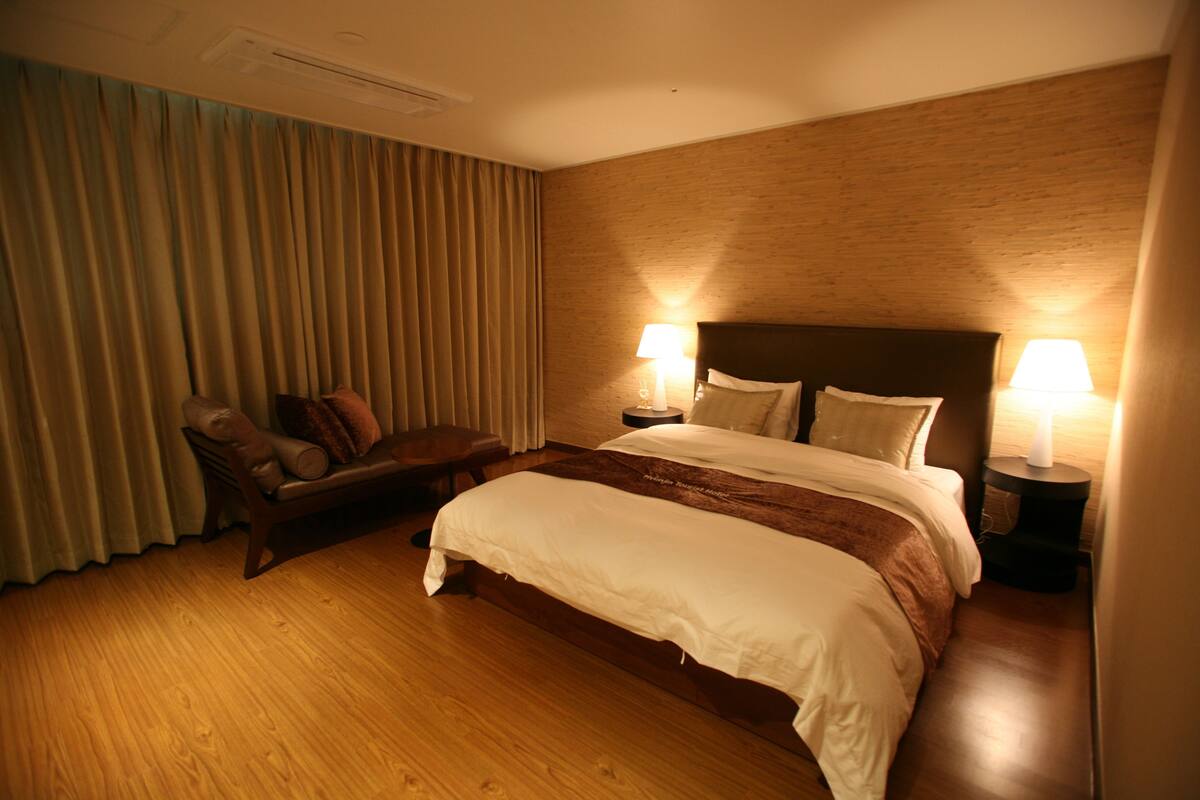 Habitación de hotel con una cama grande, dos mesitas de noche con lámparas, un cabecero de madera, suelo de madera y un ventanal con cortinas.