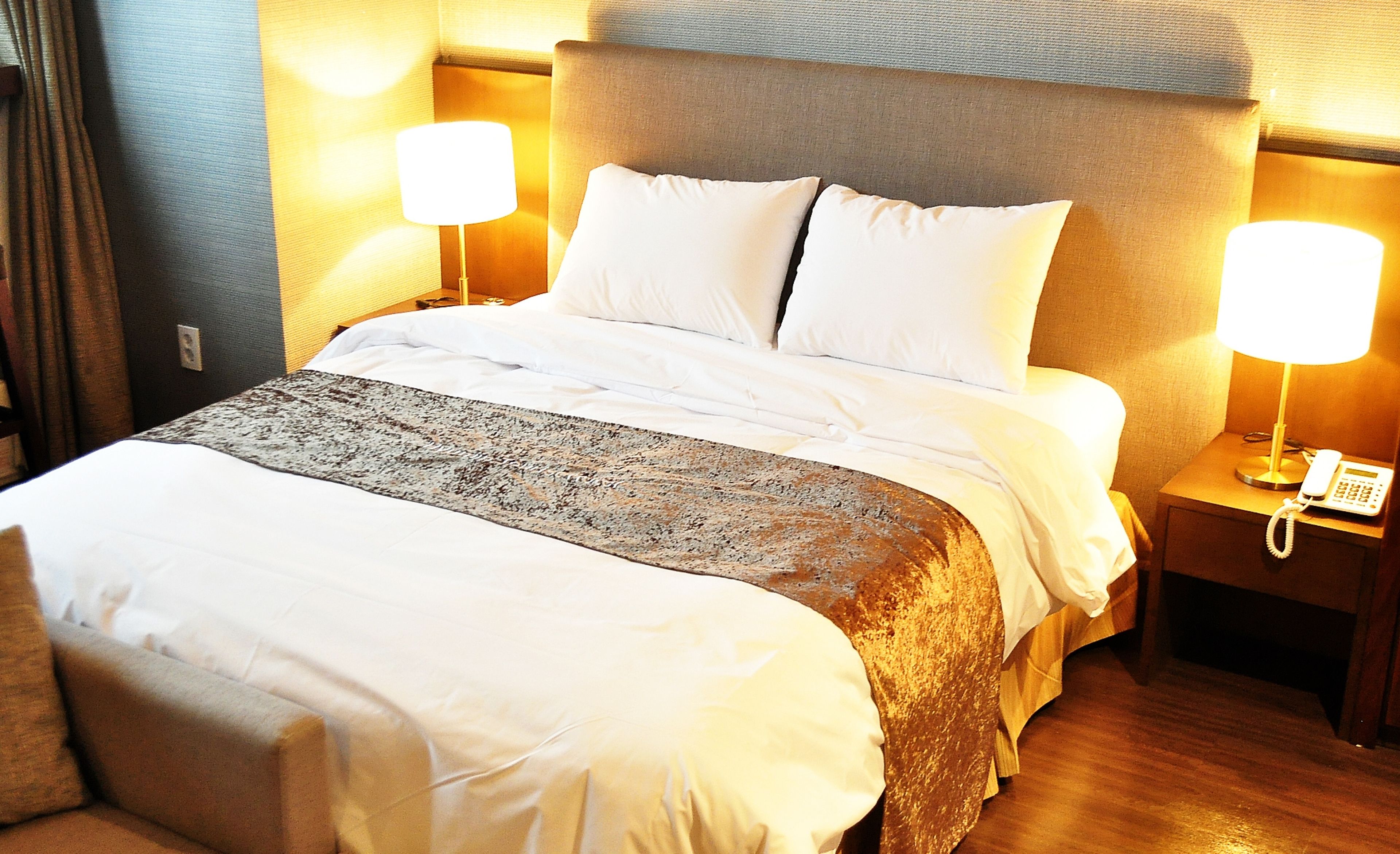 Cama bem arrumada com lençóis brancos, manta marrom e duas luminárias de apoio.