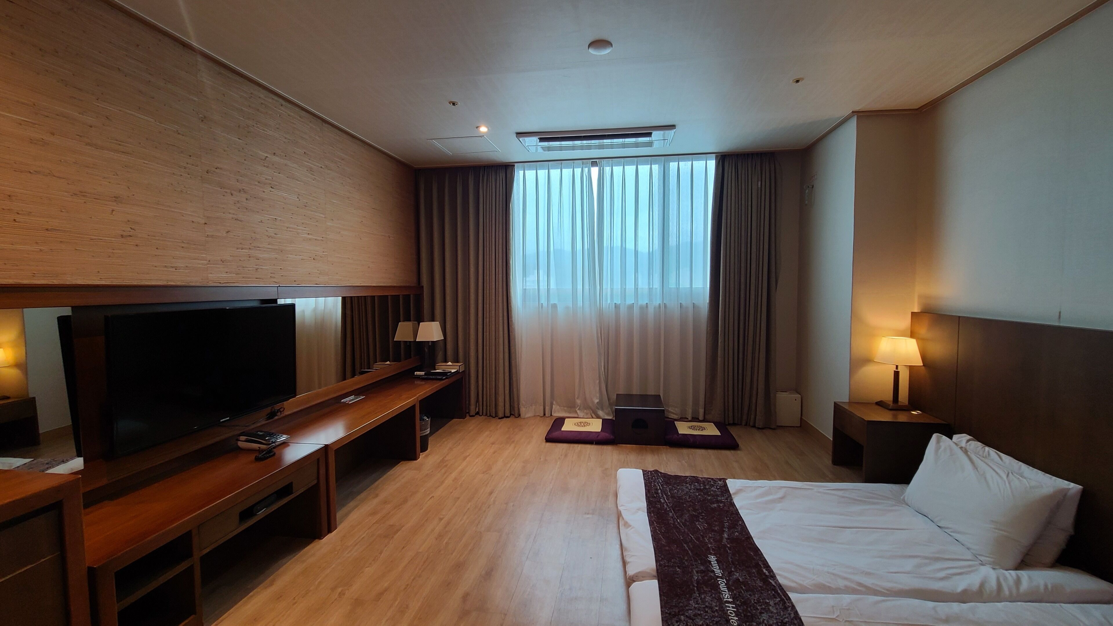 A hotel room with a large flat-screen TV, a wooden desk, a bed, and a window with curtains.