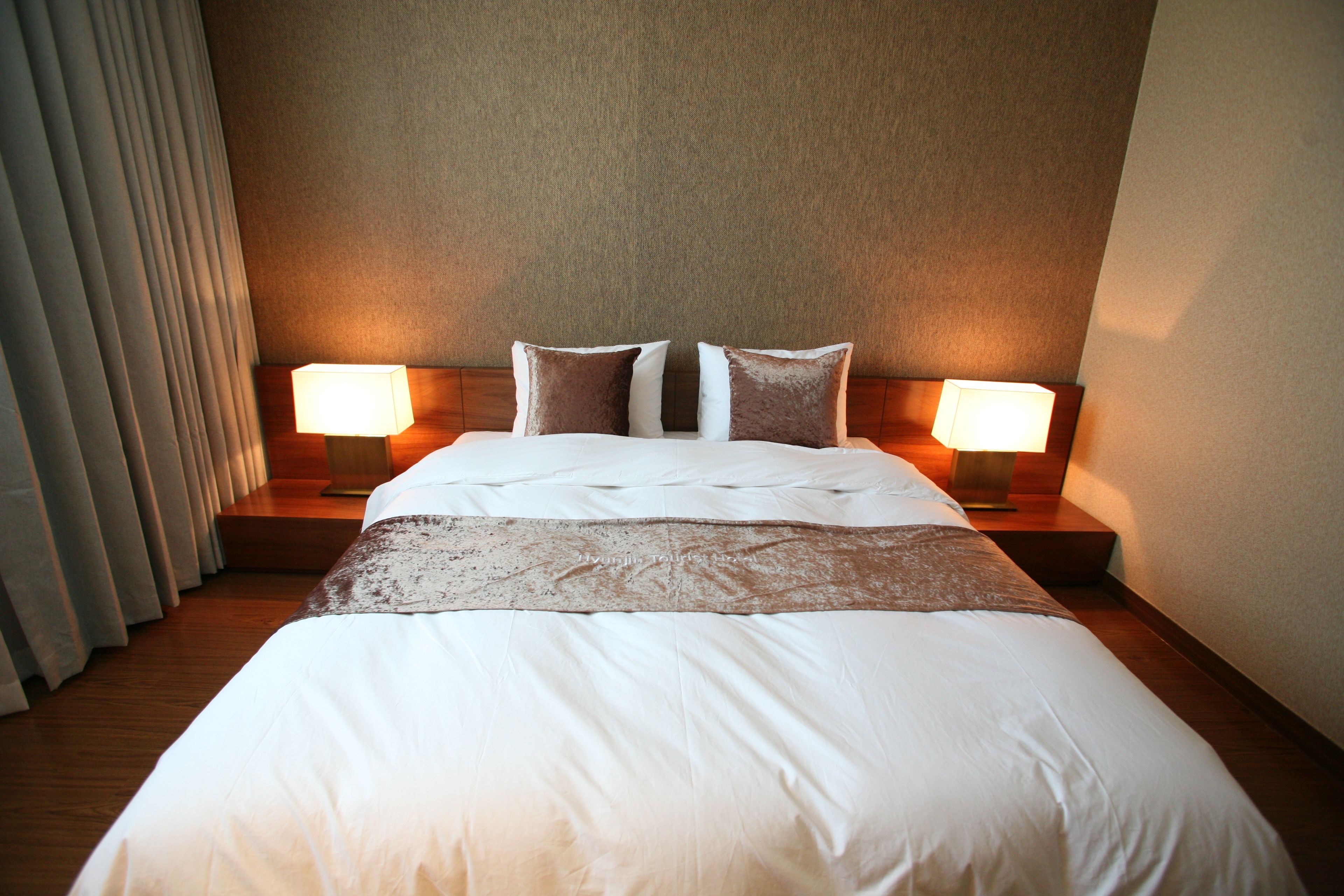Una cama bien hecha con sábanas blancas y dos almohadas, con dos lámparas de noche sobre mesitas de madera.