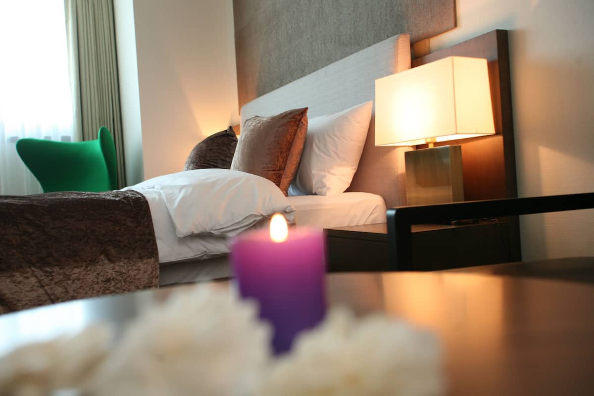 Una habitación de hotel con una cama, una mesita de noche con una lámpara, una silla verde y una vela morada.