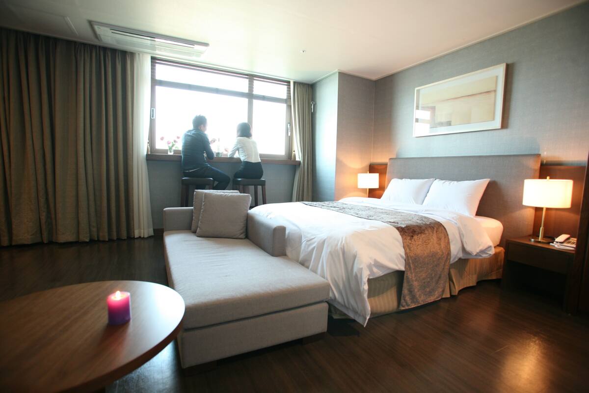 Una habitación de hotel con cama, sofá, una mesa con vela y ventana con cortinas.
