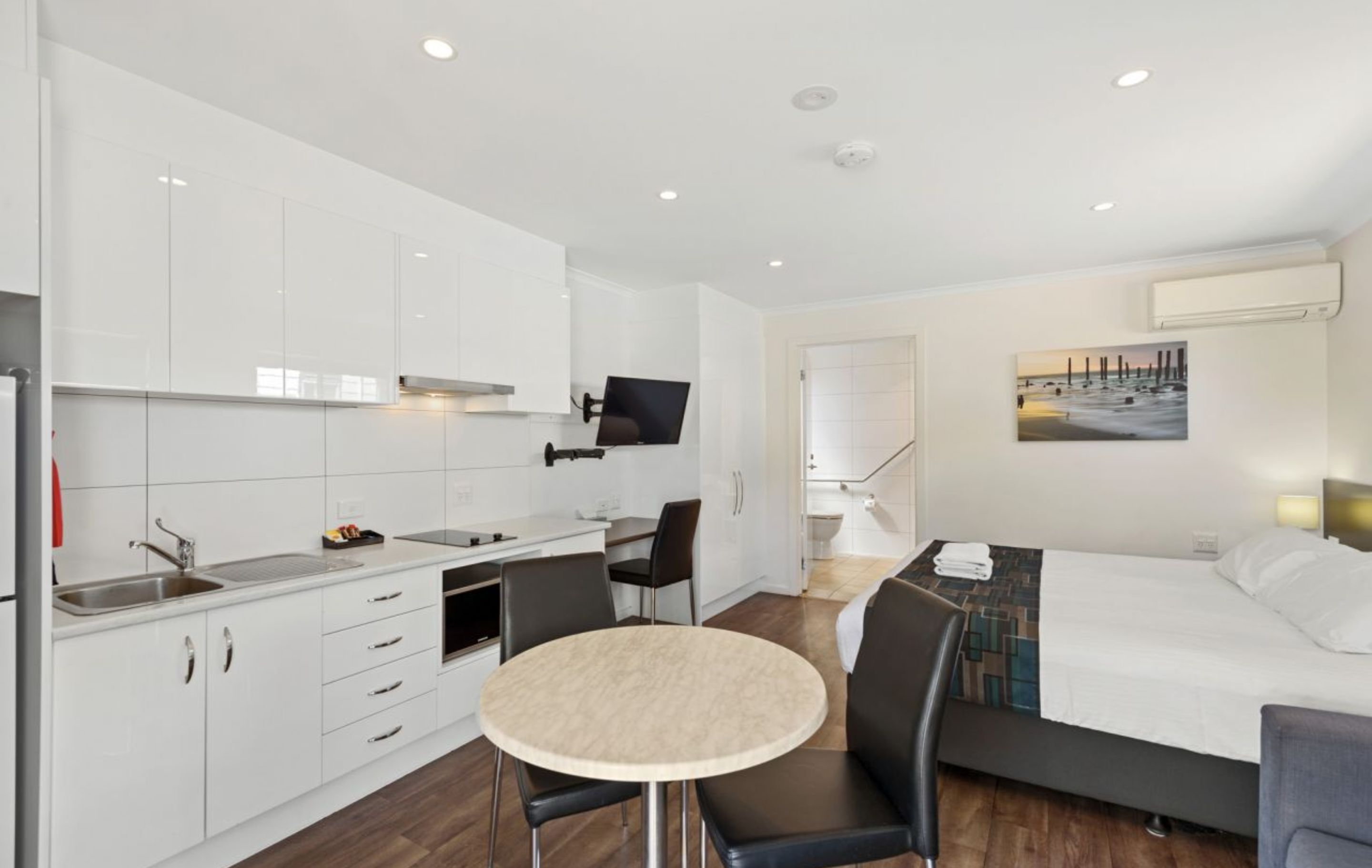 Una habitación de hotel moderna con una cama, una mesa de comedor redonda, una kitchenette y un televisor montado en la pared.