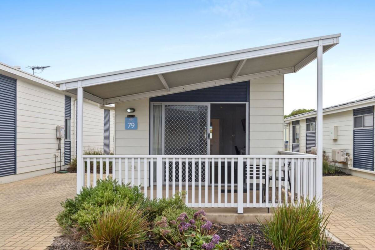 A single-story mobile home with a covered porch, a number '79' sign, and a fenced garden area with various plants.