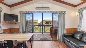 A cozy room with a wooden ceiling, a TV on a shelf, a dining table with chairs, a black leather sofa, and a sliding glass door leading to an outdoor area. - Discovery Parks - Adelaide Beachfront (Semaphore Park)