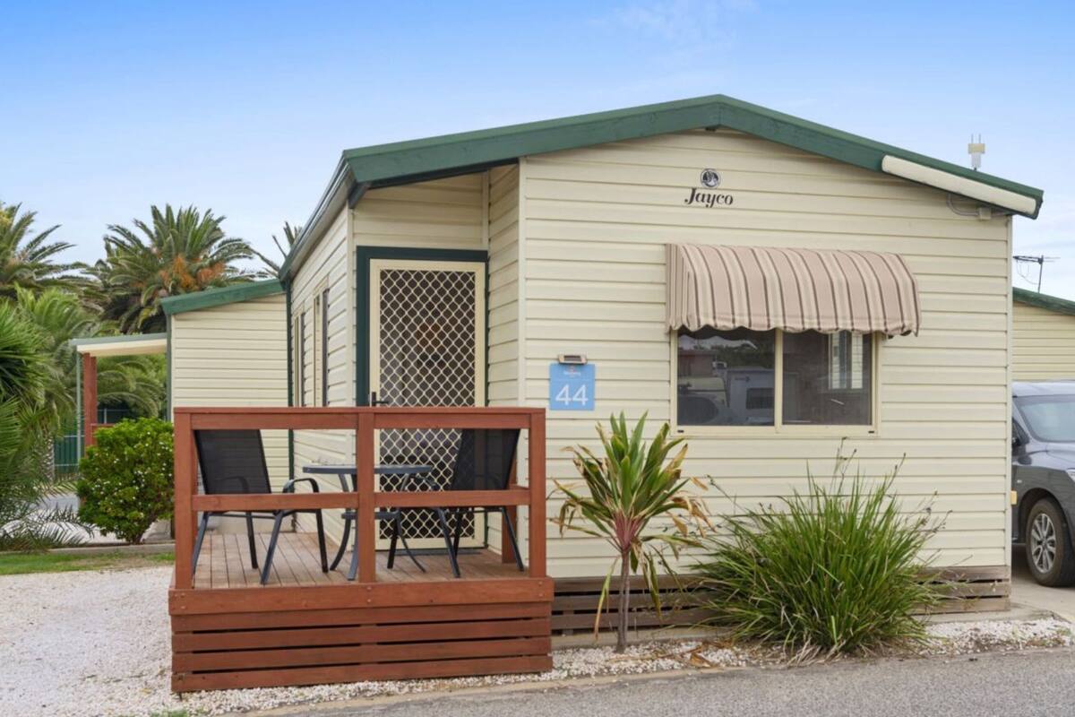 A small, single-story motel with a green roof and beige siding, featuring a porch with a table and chairs, and a number '44' sign.