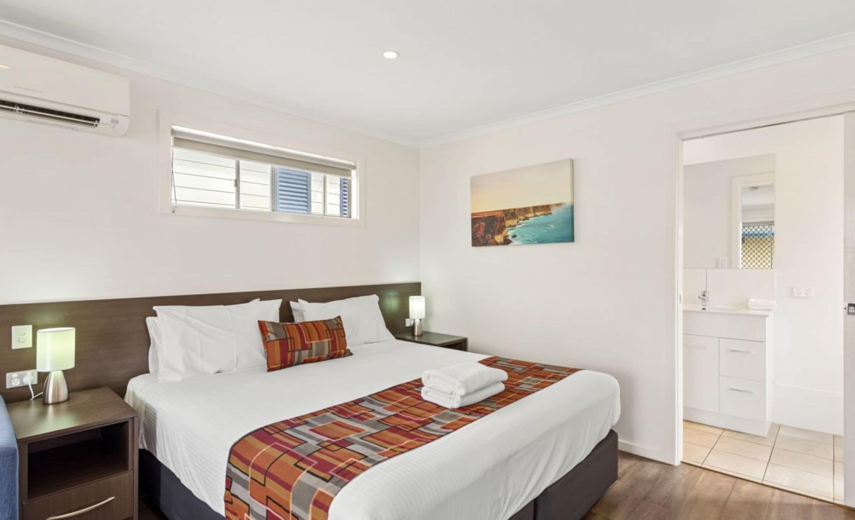 A hotel room with a bed, a nightstand, a lamp, a painting, and a bathroom visible through an open door.