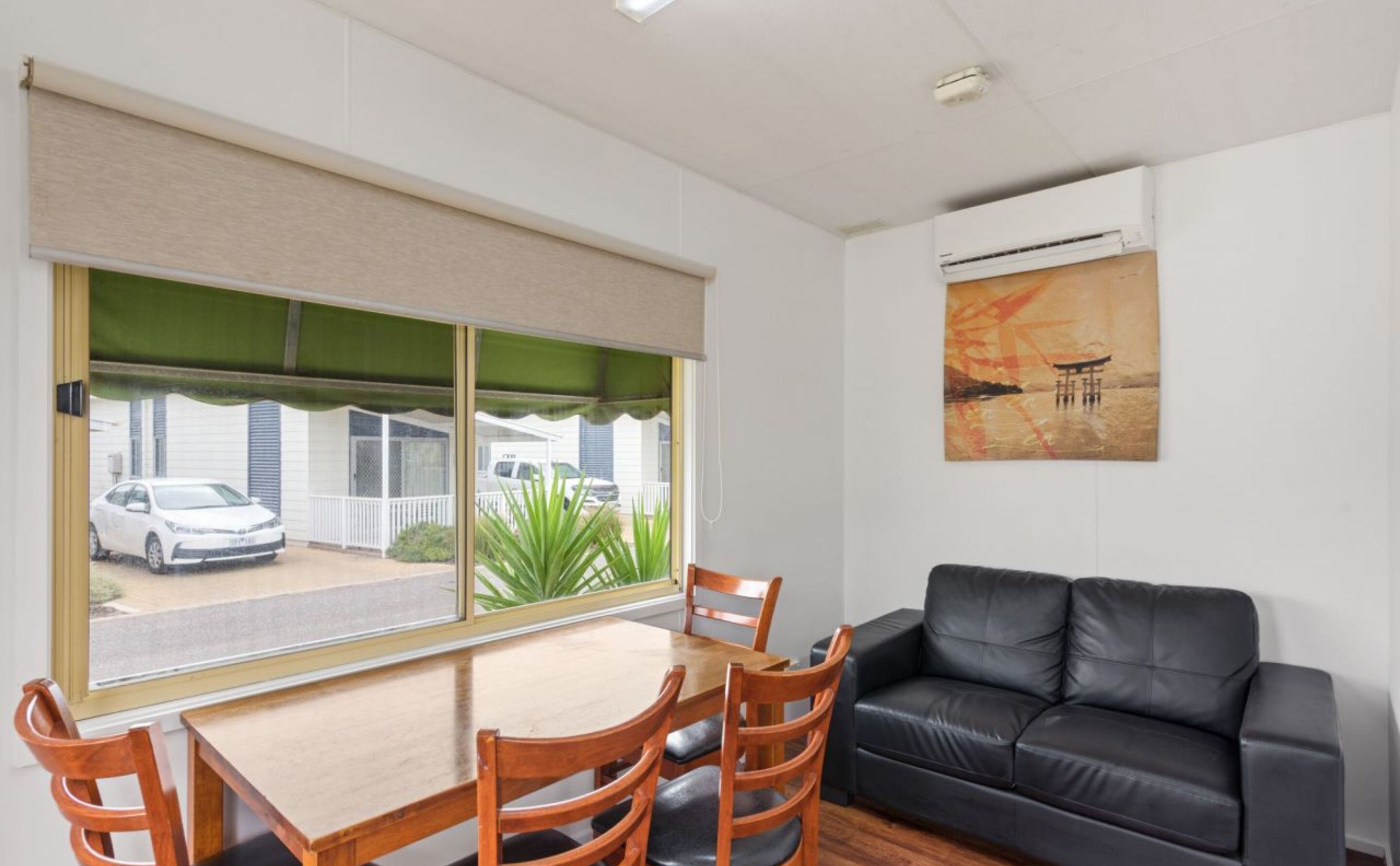 A room with a dining table and chairs, a black leather sofa, a large window with a view of a street and parked cars, and a painting on the wall.