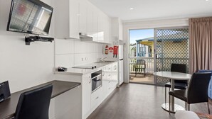 A modern kitchen with white cabinets, a built-in oven, and a flat-screen TV mounted on the wall. There is a dining area with a table and chairs, and a balcony with a view of the outside. - Discovery Parks - Adelaide Beachfront (Semaphore Park)