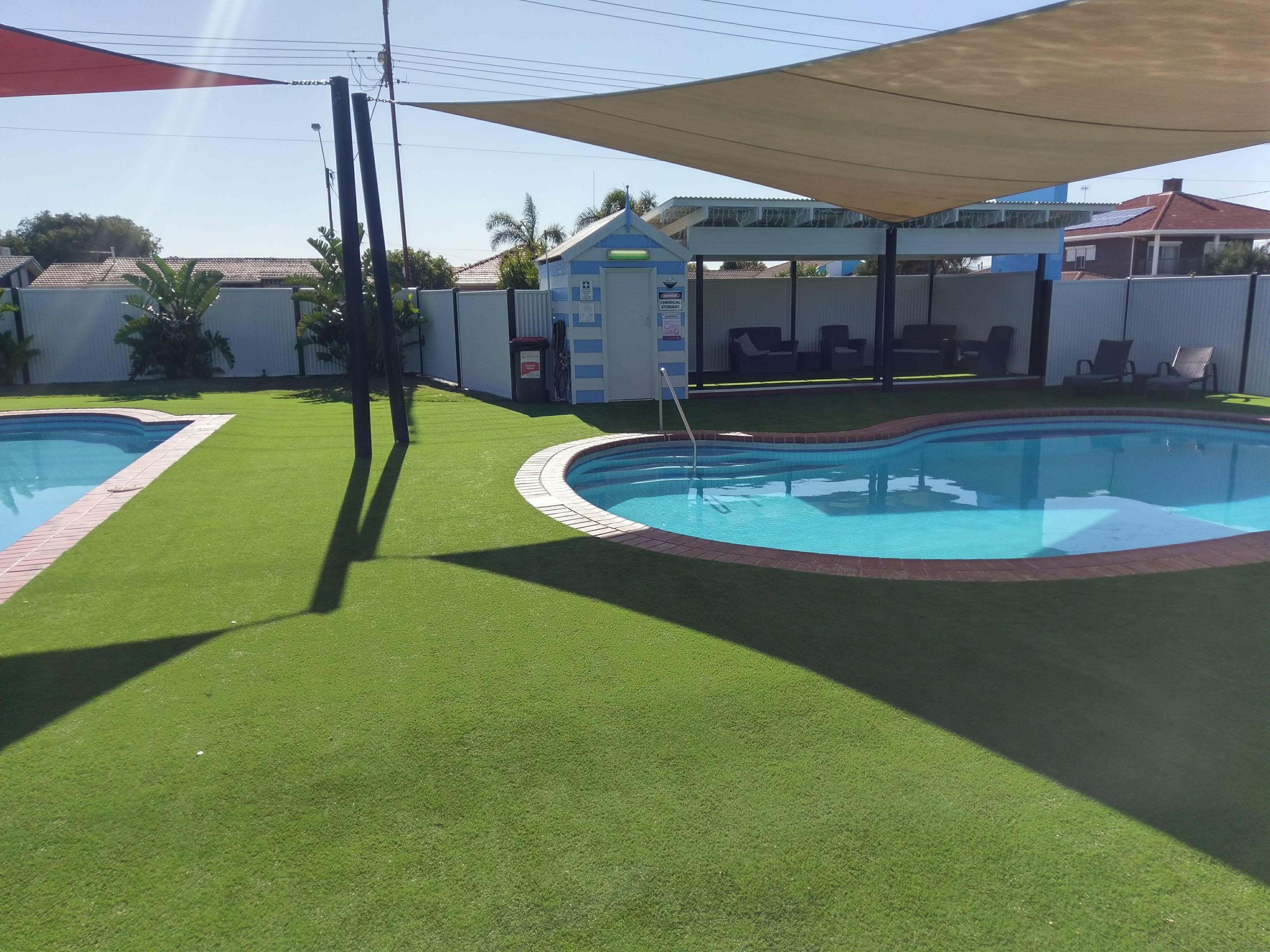A backyard with two swimming pools surrounded by artificial grass, a shaded area, and a fenced patio with outdoor furniture.
