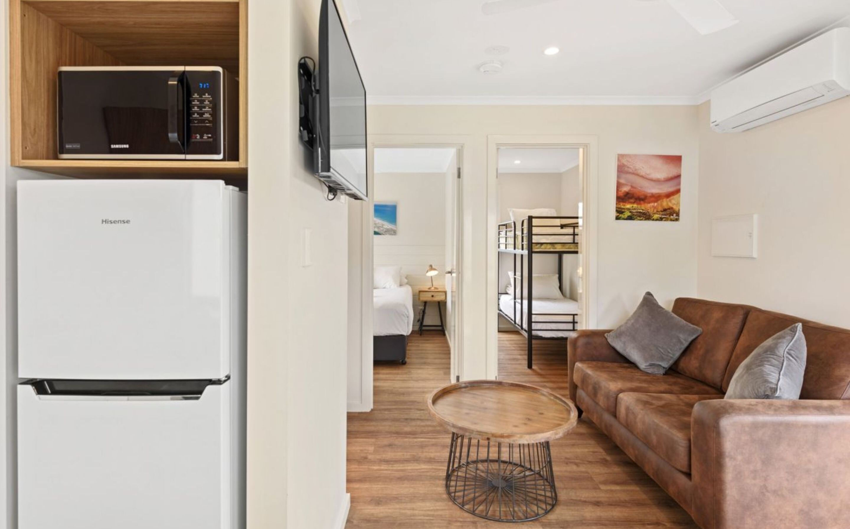 A compact living space with a white refrigerator, a microwave, a wooden dining table, a brown sofa, and a bunk bed.