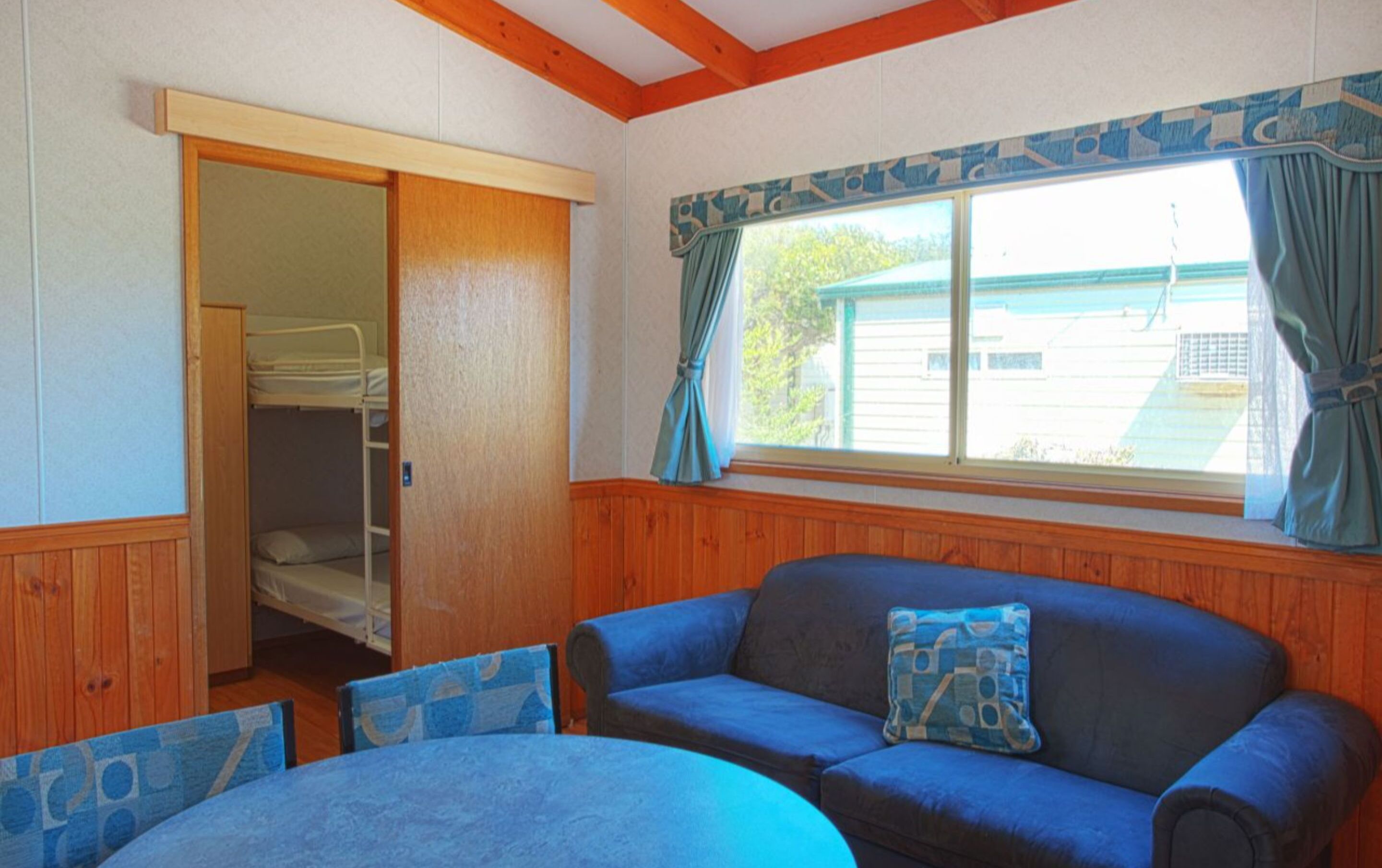 Una habitación con piso de madera, un sofá azul con un cojín a juego, una mesita redonda y una ventana con cortinas.