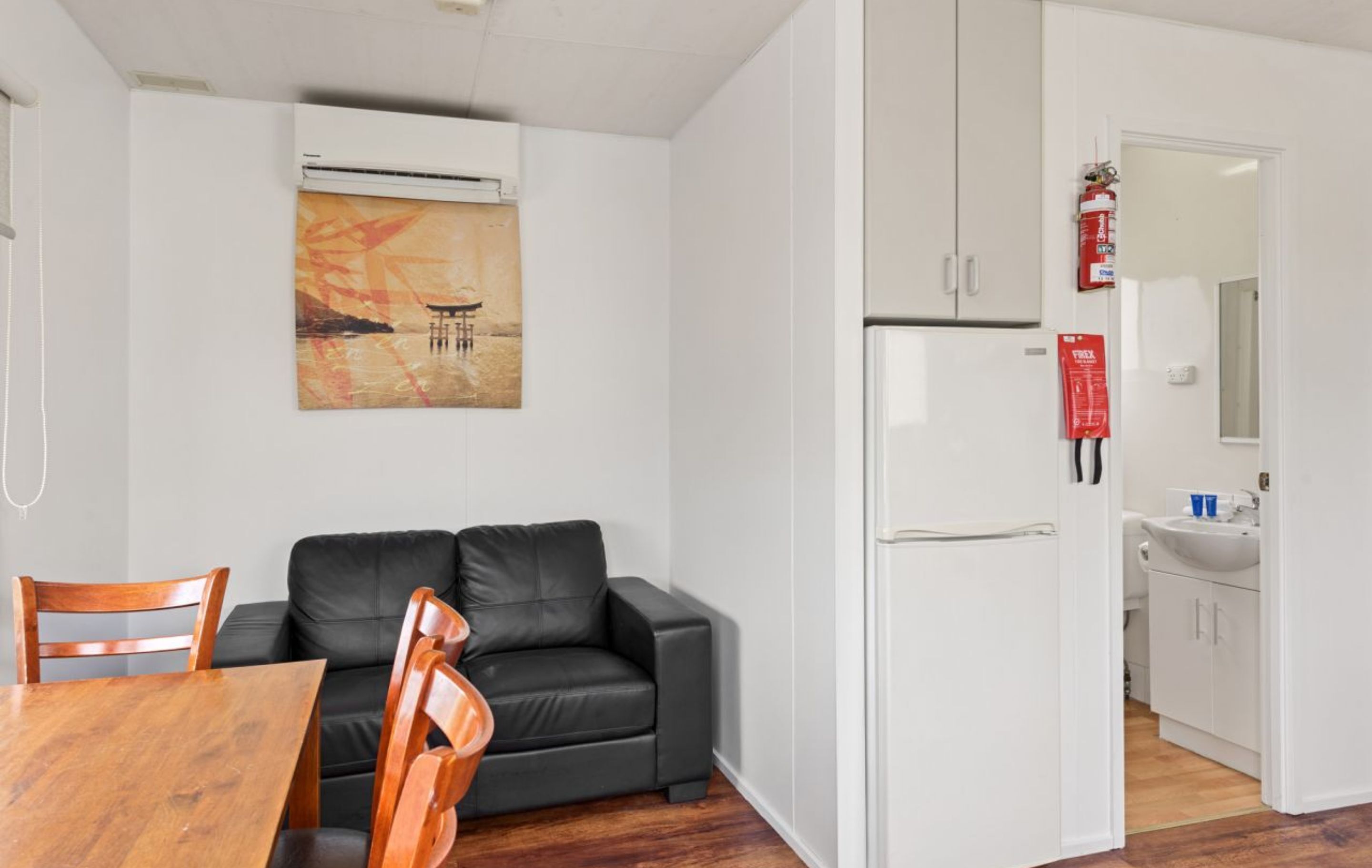 A compact living space with a dining area, a black leather sofa, a wooden dining table, and a kitchenette with a white refrigerator and sink.