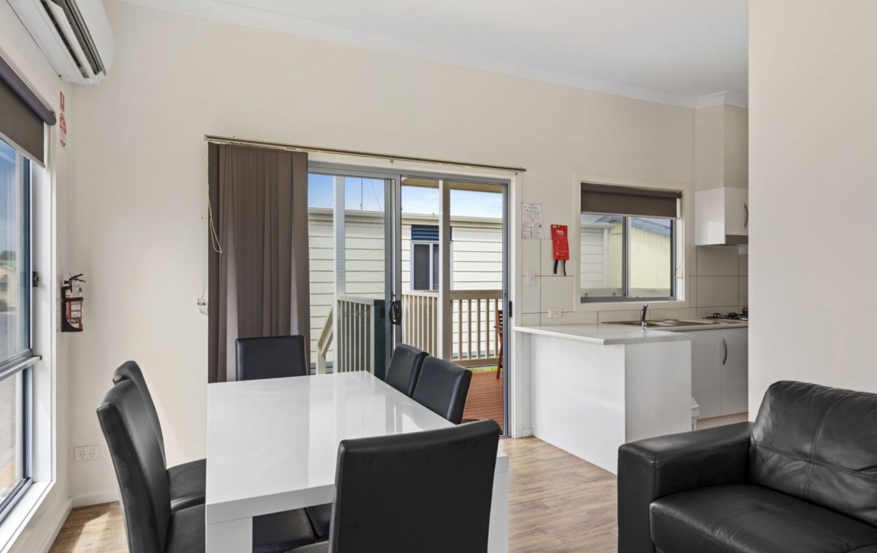 Cozinha moderna com armários brancos e uma pia, área de refeições com mesa branca e cadeiras pretas, e varanda com vista para uma casa.