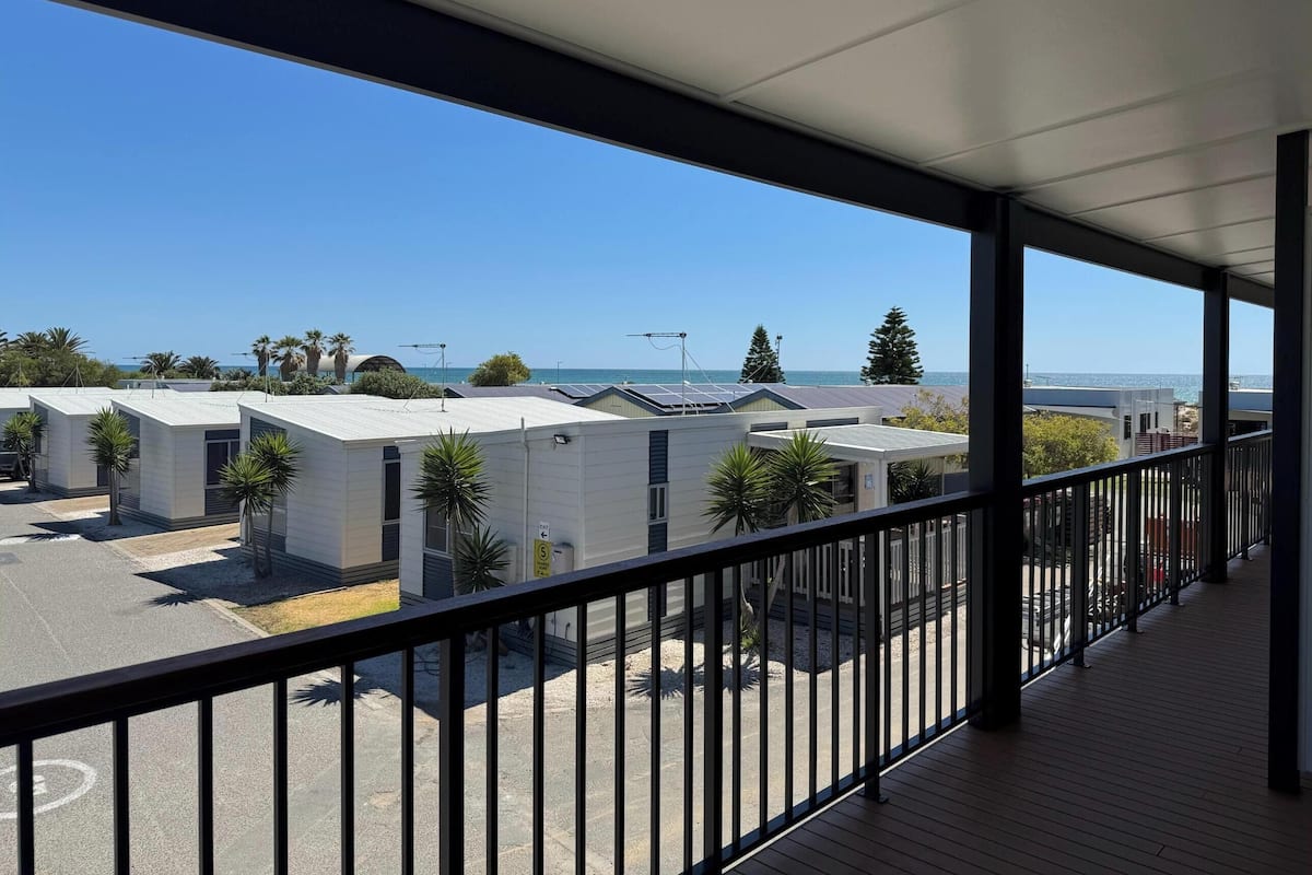 A balcony with a view of a parking area and buildings, including a row of mobile homes and a beach in the background.