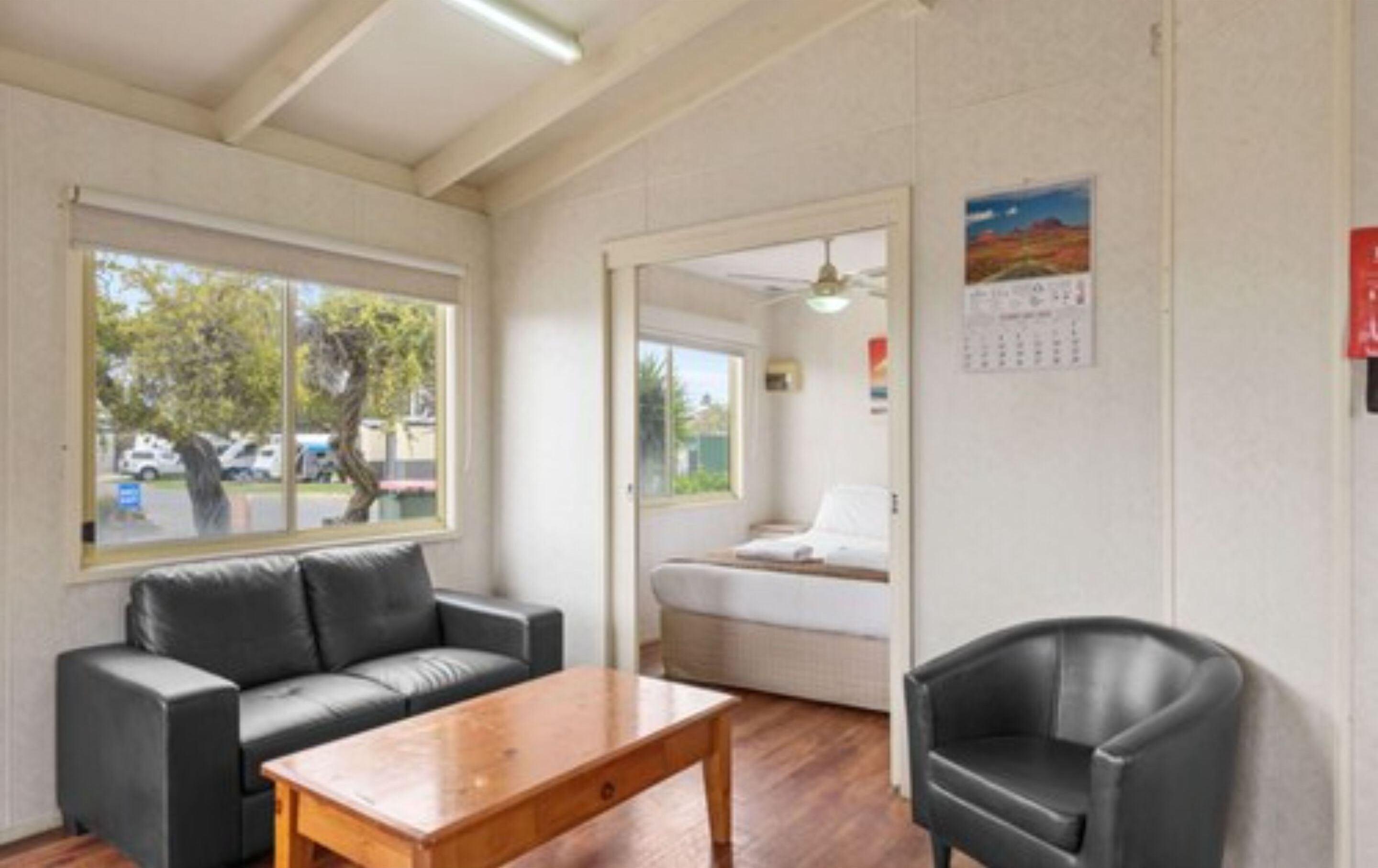 Ein Zimmer mit einer Couch, einem Holztisch und einem Sessel. Ein Fenster bietet Blick auf Bäume und parkende Autos.