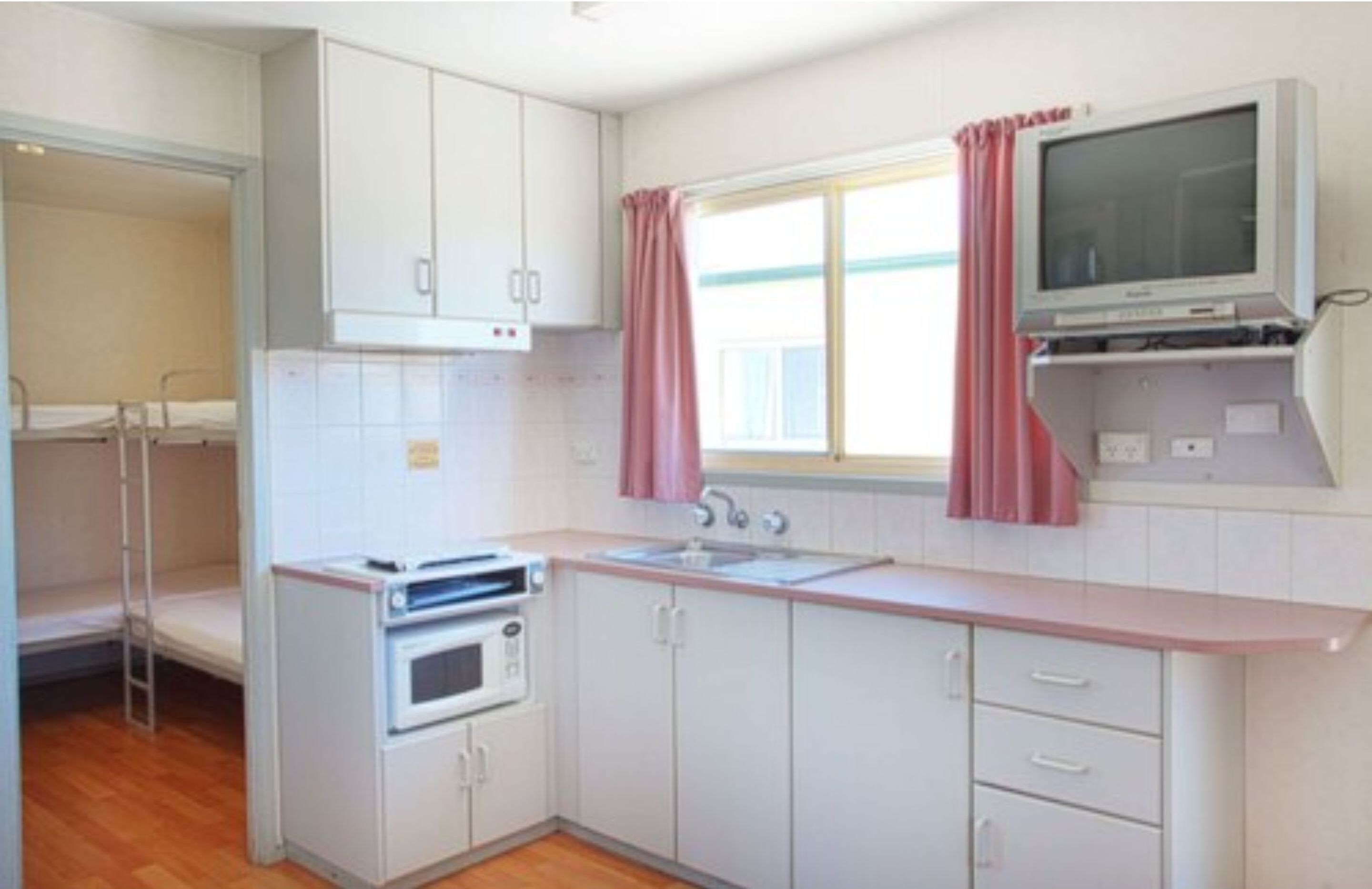 A compact kitchen with white cabinets, a microwave, a stove, a sink, and a television mounted above the counter.