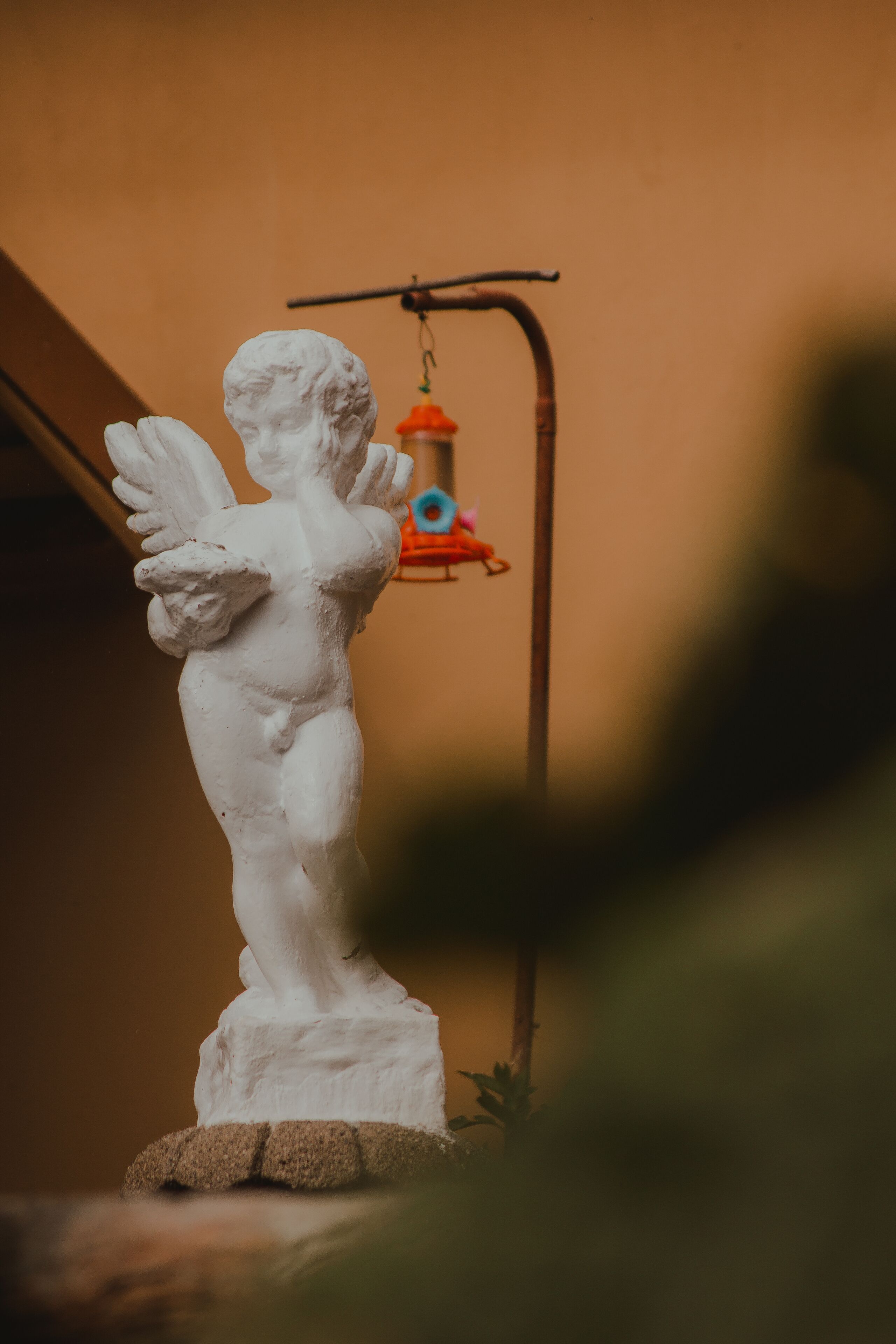 Une statue blanche représentant une figure ailée, posée sur un socle, avec un abreuvoir à oiseaux en arrière-plan.
