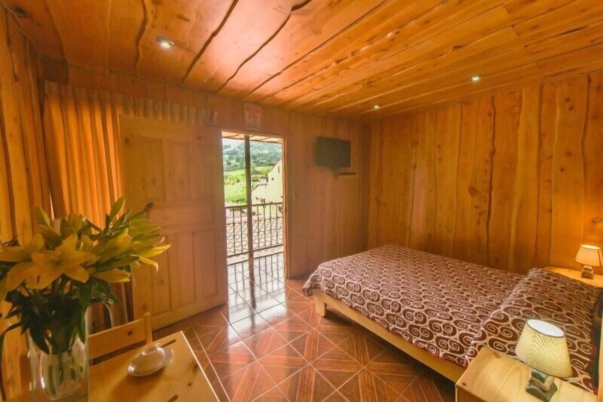Ein Zimmer aus Holz mit einem Bett, einem Tisch mit Blumenvase und Blick ins Freie durch eine Tür.