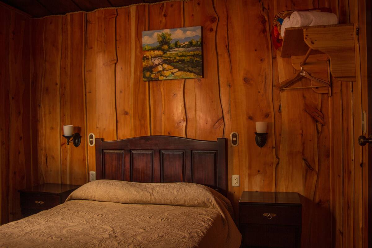 Ein Zimmer mit Holzwänden, einem Bett, zwei Wandleuchten, einem Gemälde und einer Holzkommode.
