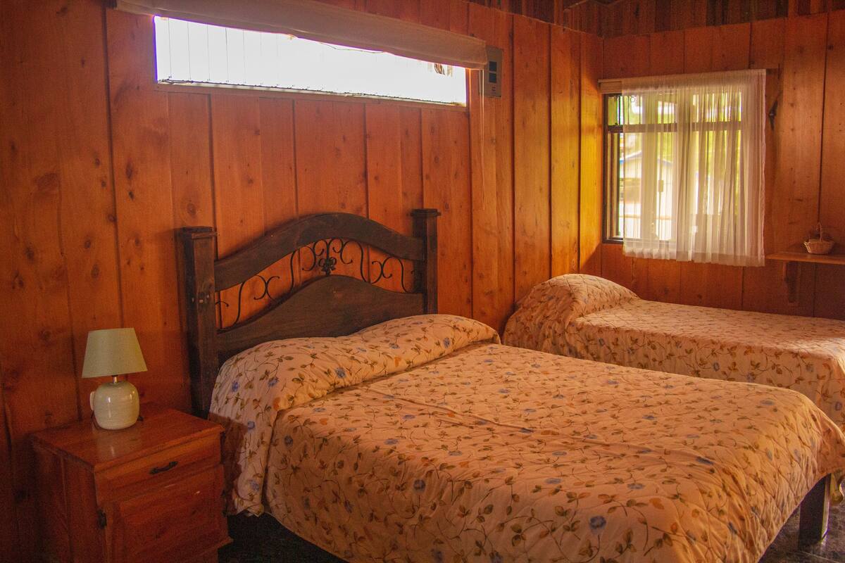 Ein Schlafzimmer in einer Holzhütte mit zwei Betten, einem Nachttisch und einem Fenster mit Vorhängen.