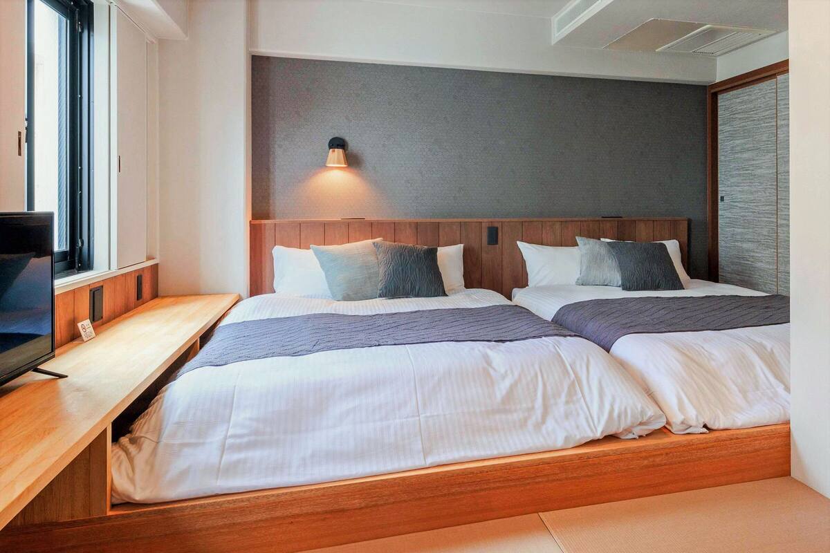 Une chambre d’hôtel avec deux lits, une tête de lit en bois, une lampe fixée au mur et un téléviseur posé sur un meuble en bois.