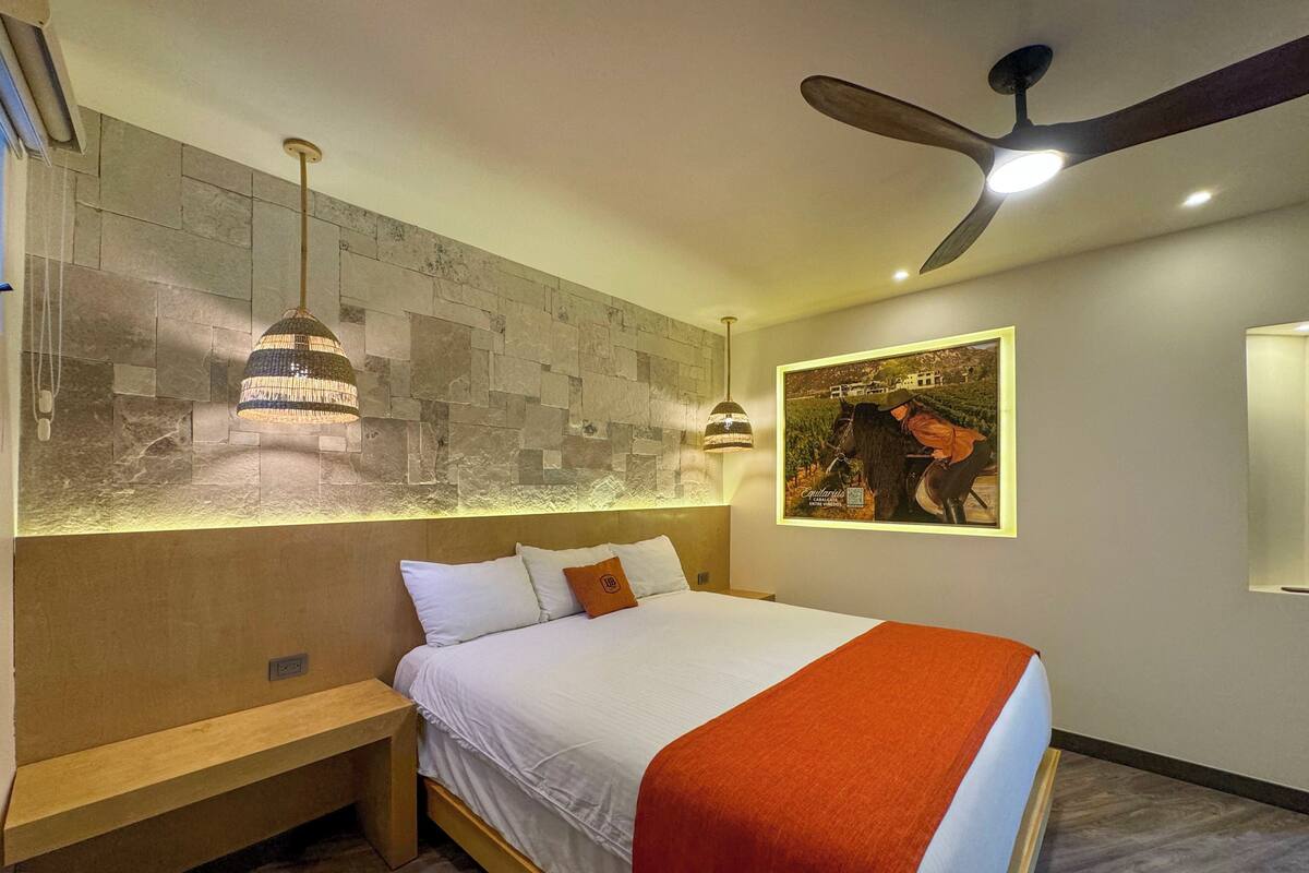 A modern bedroom with a bed, bench, pendant lights, a ceiling fan, and a wall-mounted artwork.