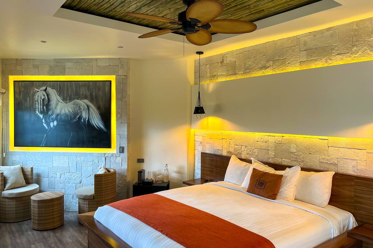 A bedroom with a large bed, a mounted painting, wicker furniture, and a stone wall.