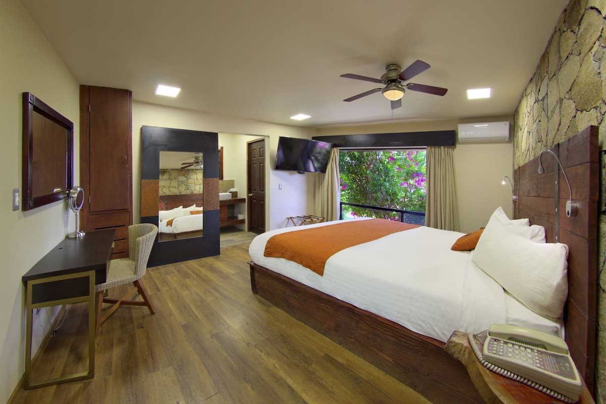 A bedroom with a large bed, a desk, a chair, a TV, and a balcony with a view of trees.