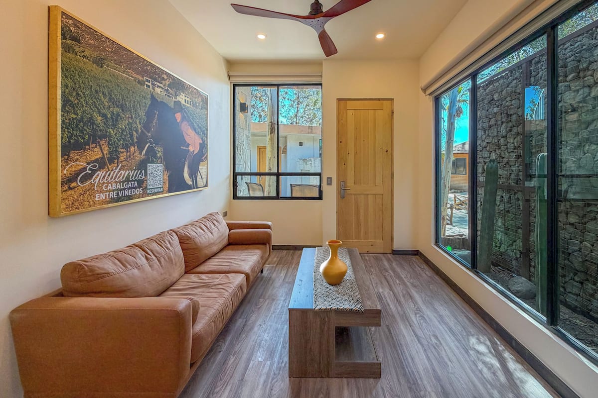 A living room with a brown leather sofa, a wooden coffee table, a ceiling fan, and a large window with a view of a stone wall and greenery.
