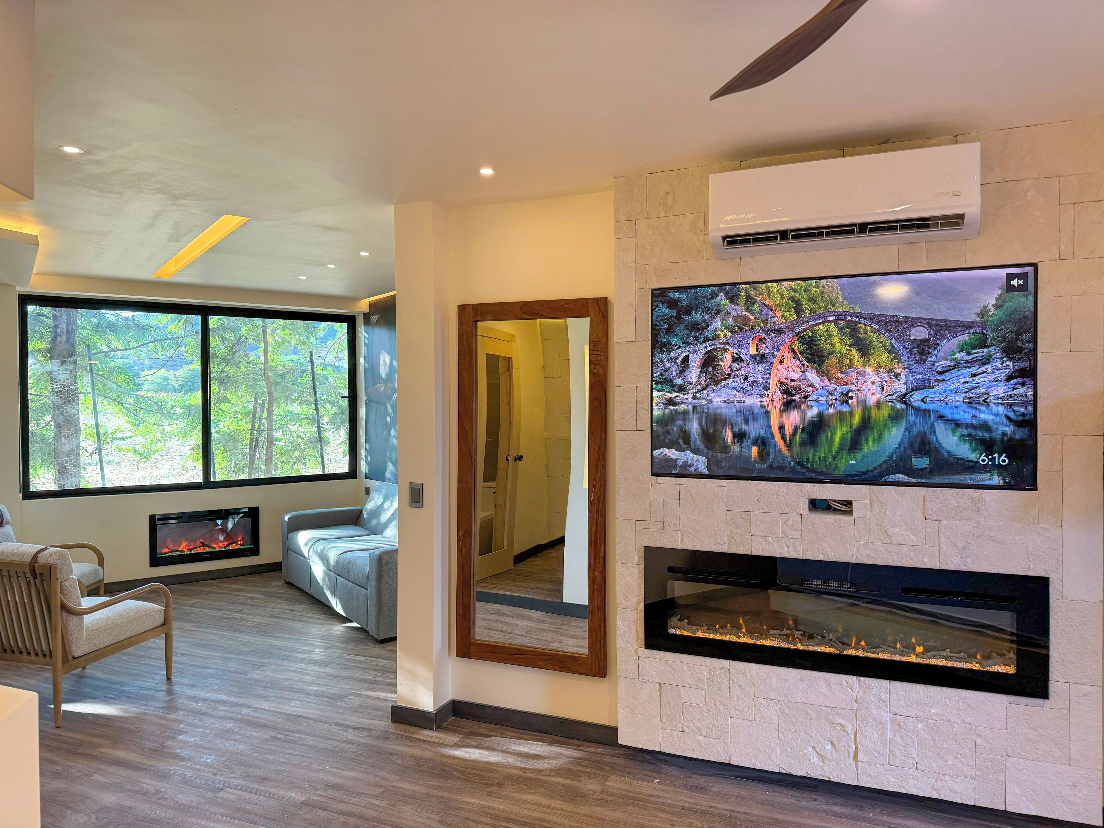 A modern living room with a large TV, fireplace, and large windows offering a view of trees.