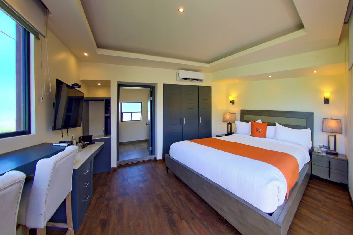 A hotel room with a large bed, a desk, a chair, a TV, and an air conditioning unit.