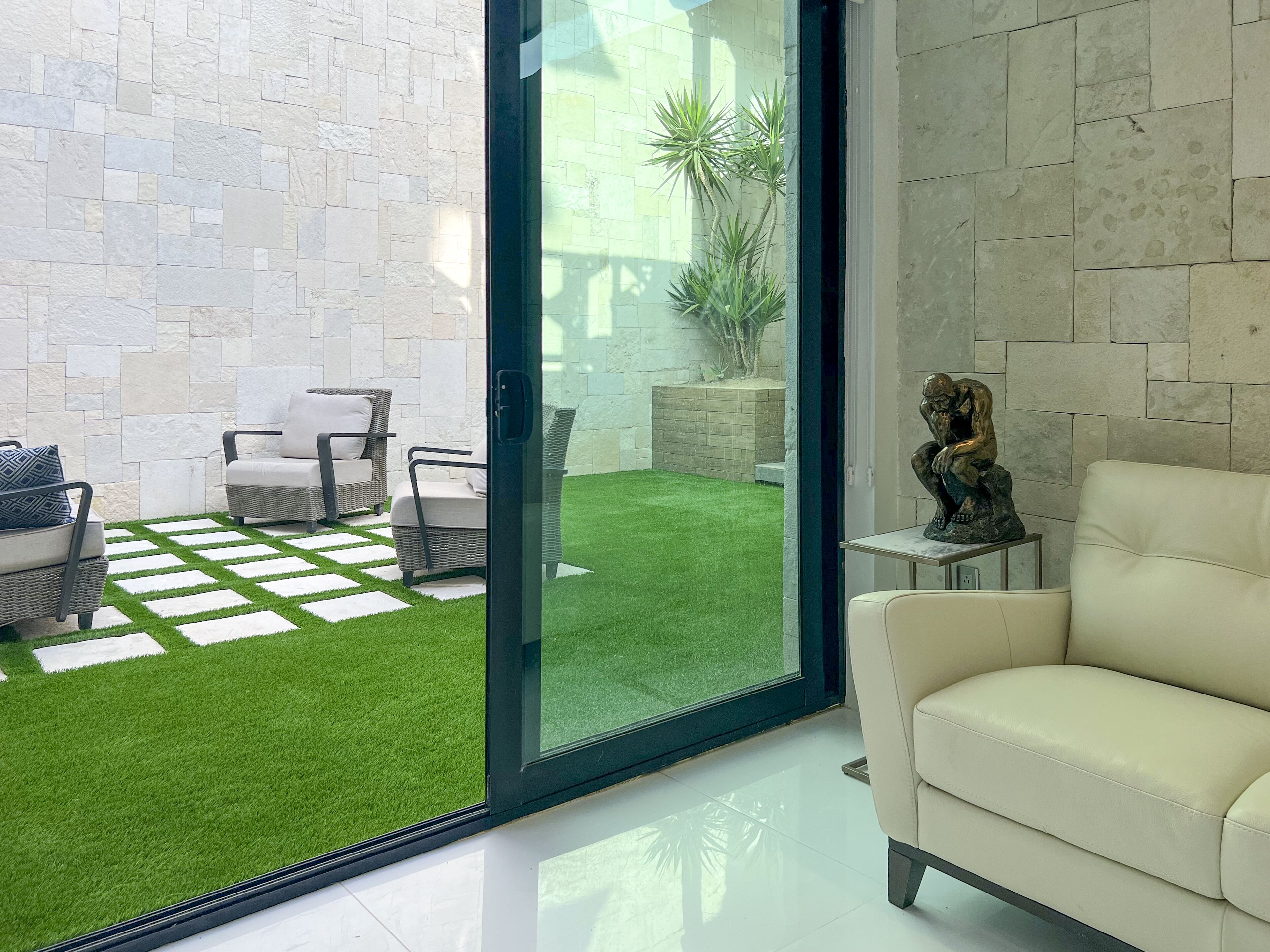A modern living room with a white sofa, a glass door leading to a patio with artificial grass and outdoor furniture.
