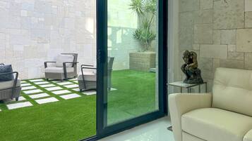 A modern living room with a white sofa, a glass door leading to a patio with artificial grass and outdoor furniture.