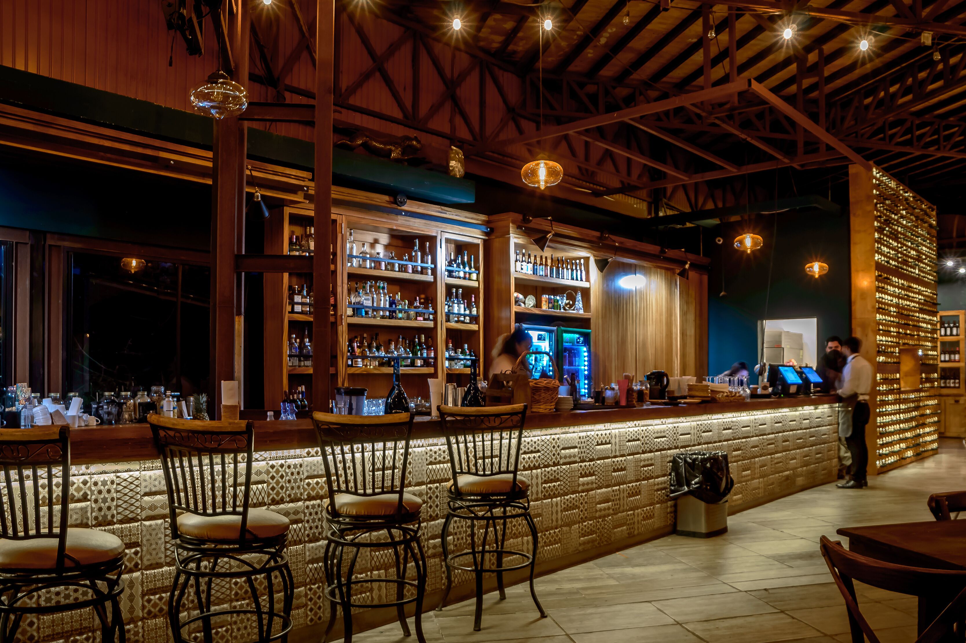 Zona de bar con estantes de madera llenos de botellas, barra y taburetes.