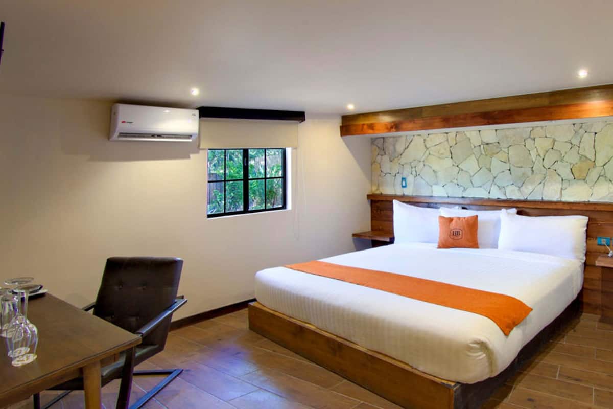 A hotel room with a bed, a desk with chairs, an air conditioning unit, a window, and a stone wall.