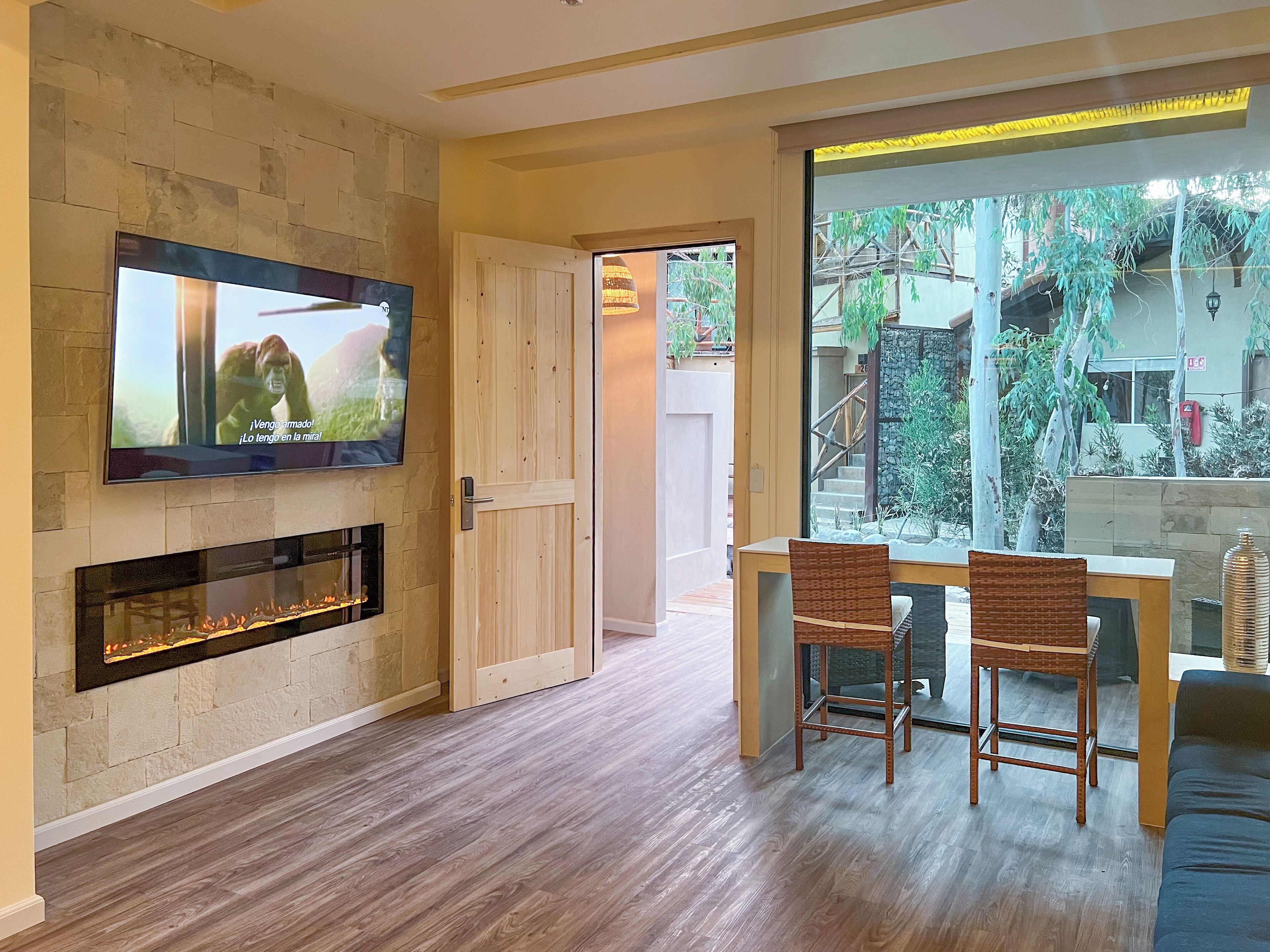 A modern living room with a fireplace, a TV mounted on a stone wall, and a dining area with wicker chairs.