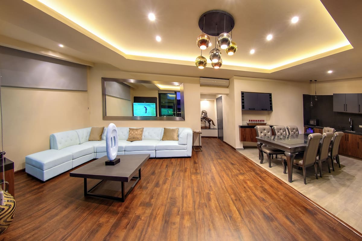 A modern living room with a sectional sofa, a dining table with chairs, a TV mounted on the wall, and a kitchen area with cabinets and a refrigerator.