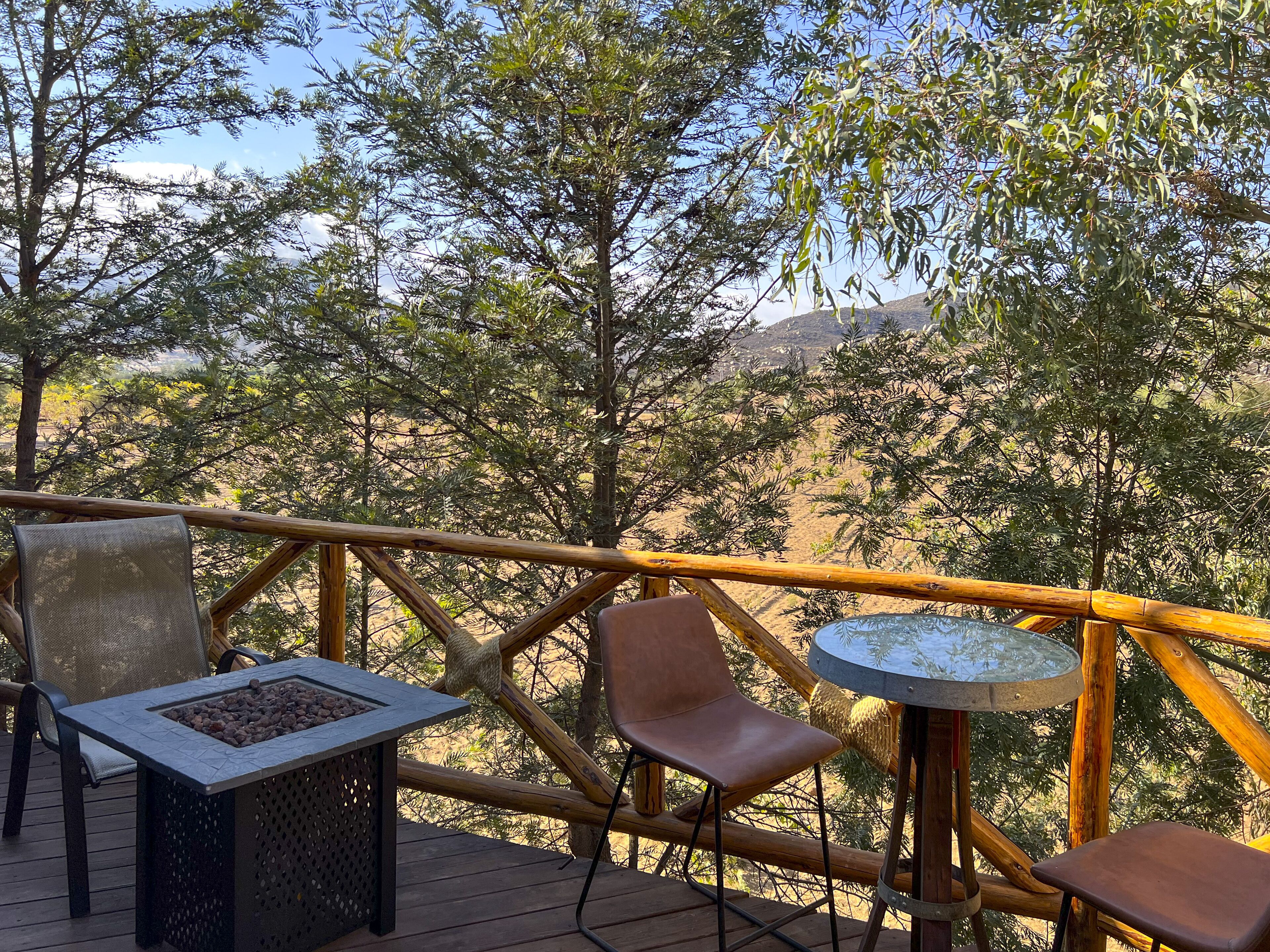 Une terrasse en bois, agrémentée d’une table et de chaises, entourée d’une clôture en bambou, et offrant une vue sur un paysage désertique à la végétation clairsemée.