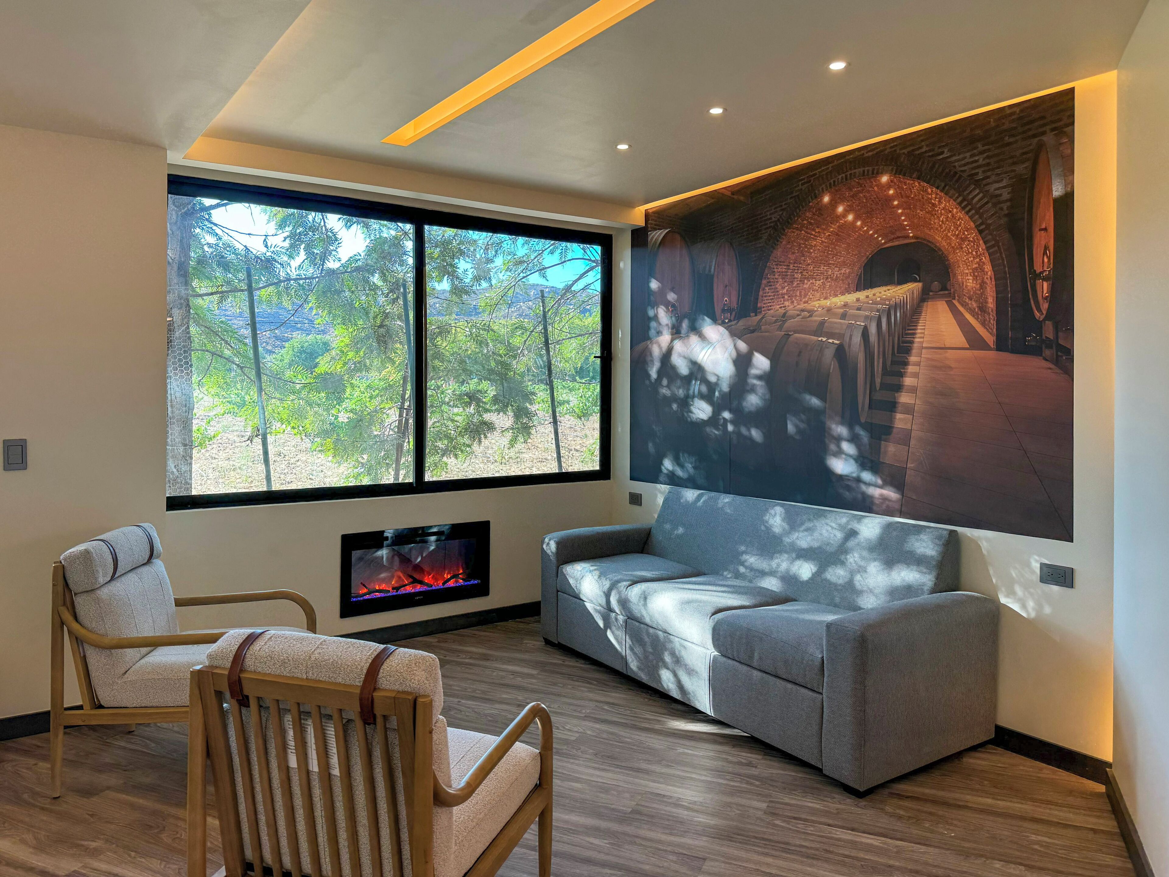 A modern living room with a fireplace, a large window with a view of trees, and a wall-mounted projector displaying a scene.