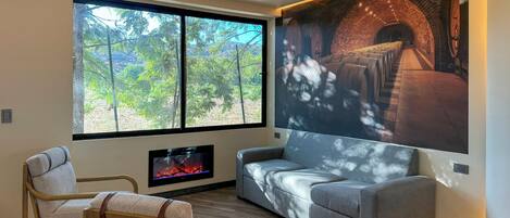 A modern living room with a fireplace, a large window with a view of trees, and a wall-mounted projector displaying a scene.